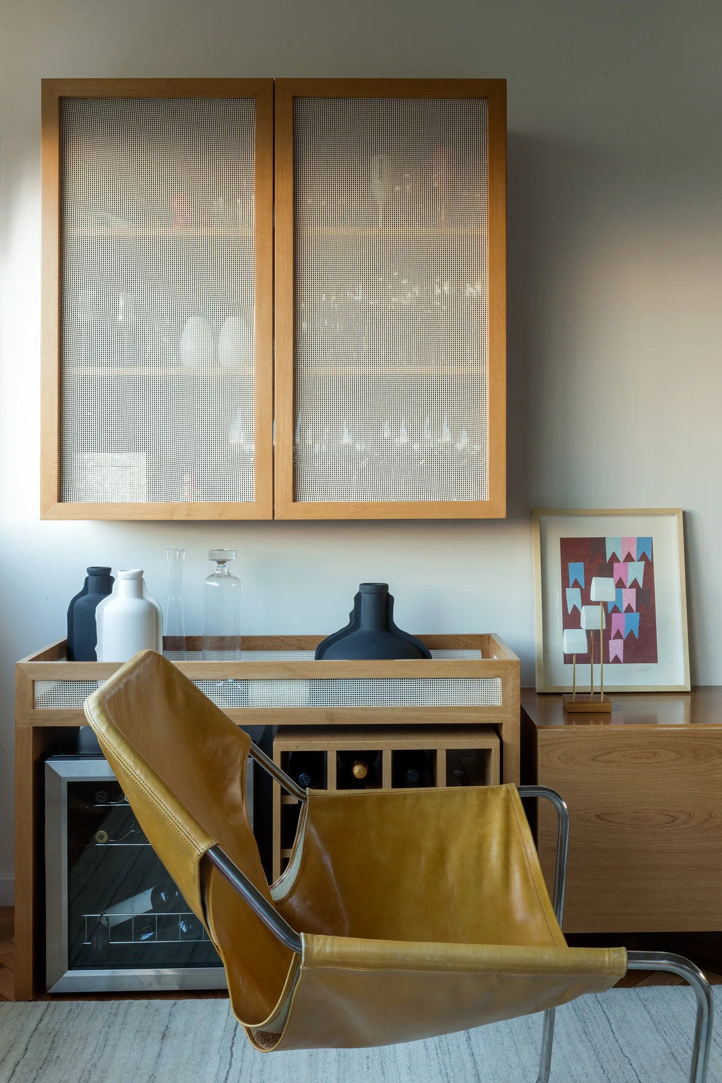 A light wood console with a built-in wine cooler and rack stands below a wall-mounted mesh cabinet next to a mustard leather lounge chair.