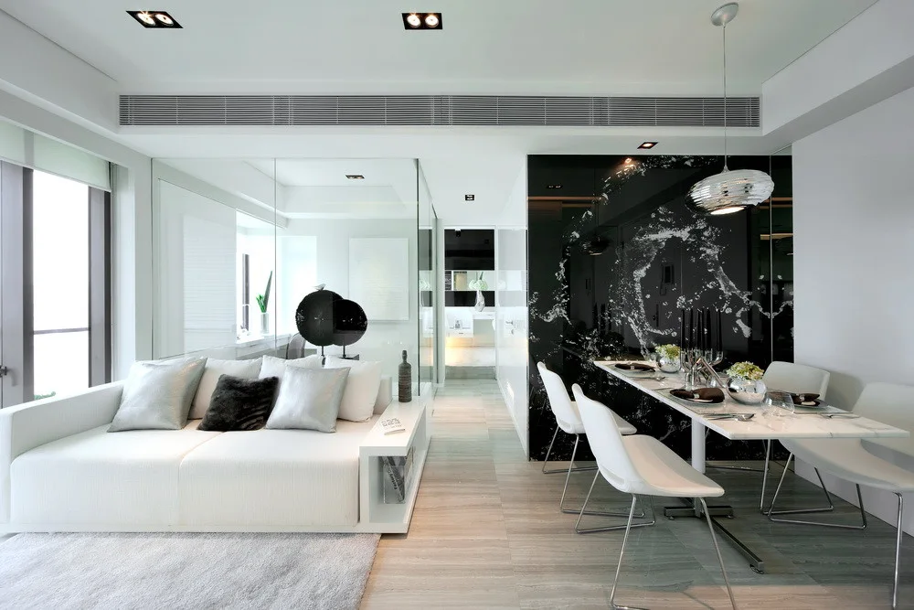 A modern open-concept space featuring a white sectional sofa next to a glass partition, opposite a marble dining table and a glossy black veined wall.