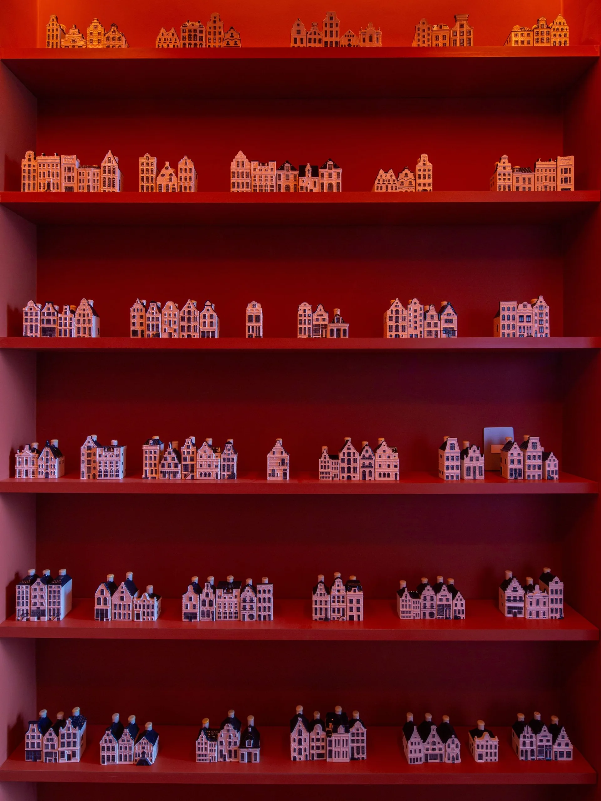 Rows of miniature ceramic canal houses are displayed on five tiers of a matte red painted shelving unit.