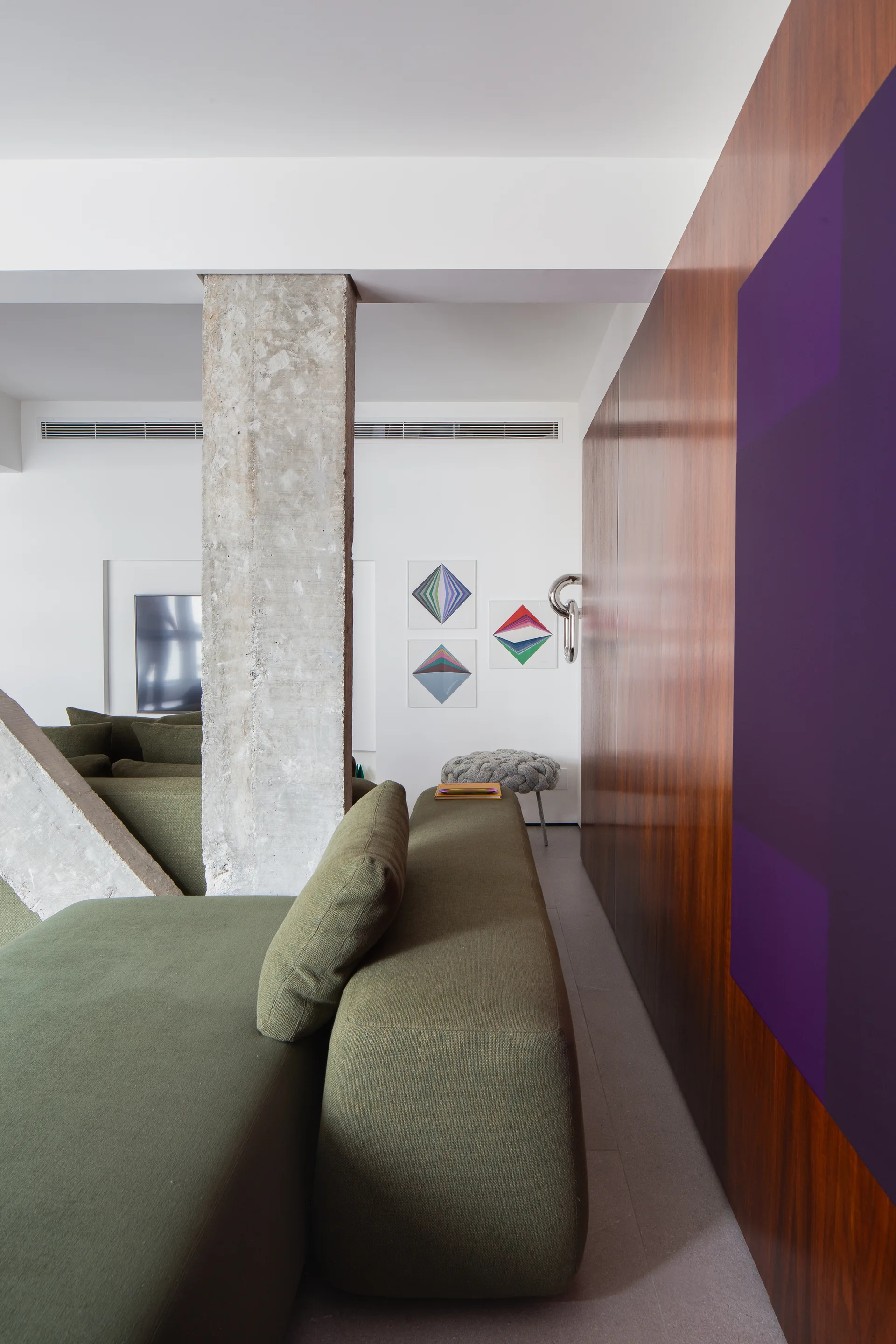 A modern living room features an olive green modular sofa, a raw concrete column, and warm wood wall paneling with a purple accent.