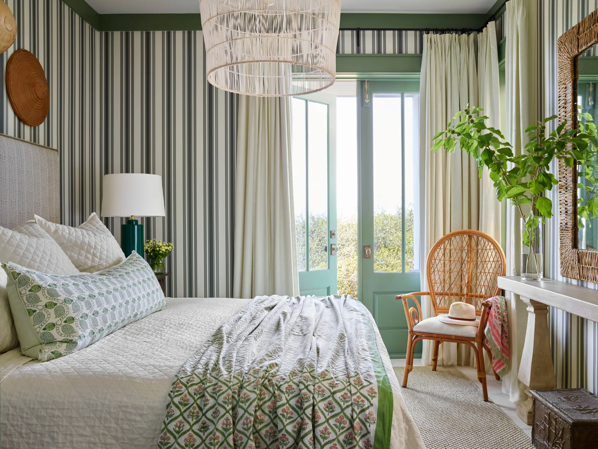 A bedroom with striped wallpaper, a bed with green patterned pillows, a rattan chair, and open green French doors leading outside.