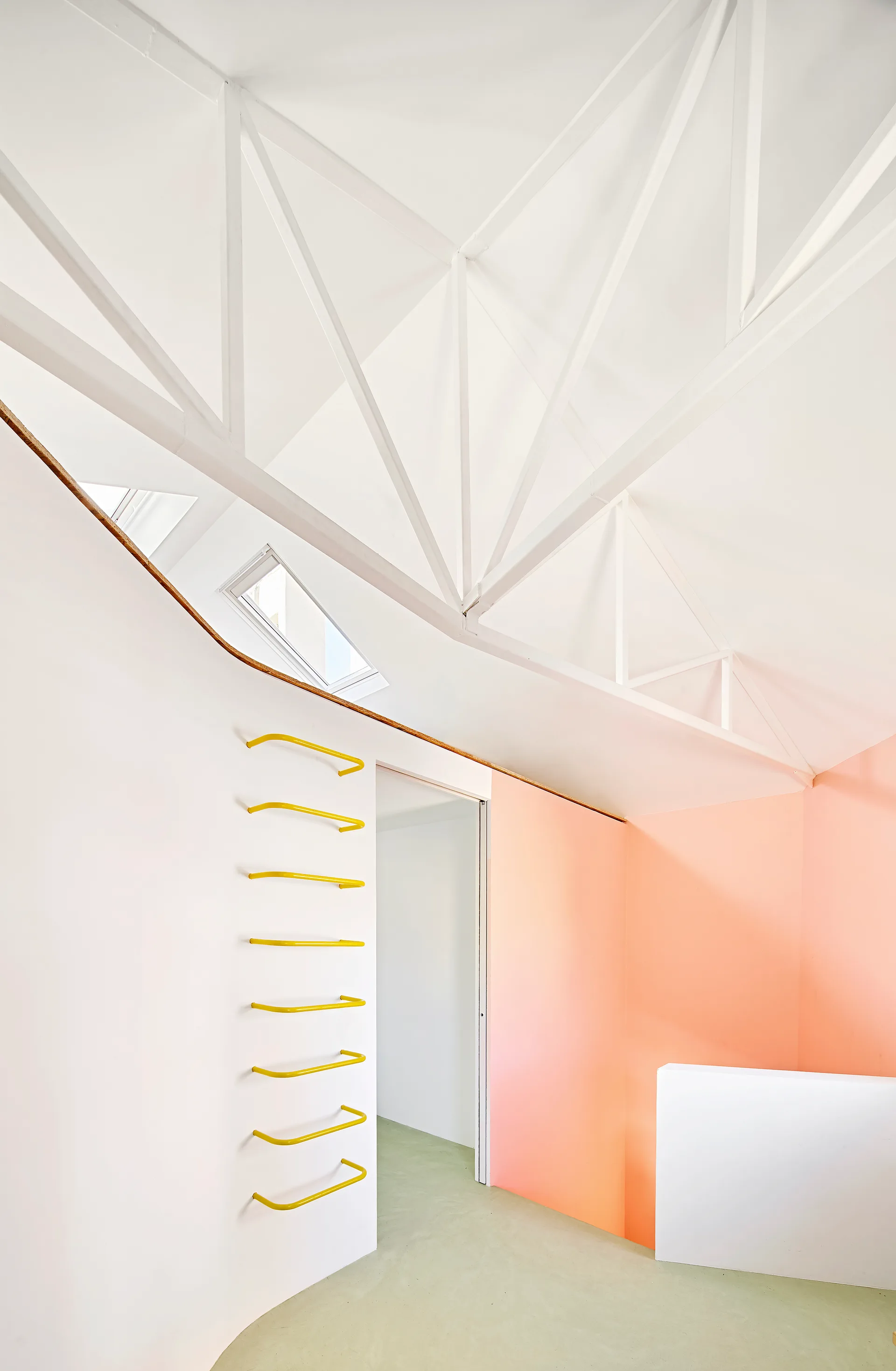An interior space features white exposed ceiling trusses, a bright yellow ladder on a curved wall, and a peach partition with a doorway.