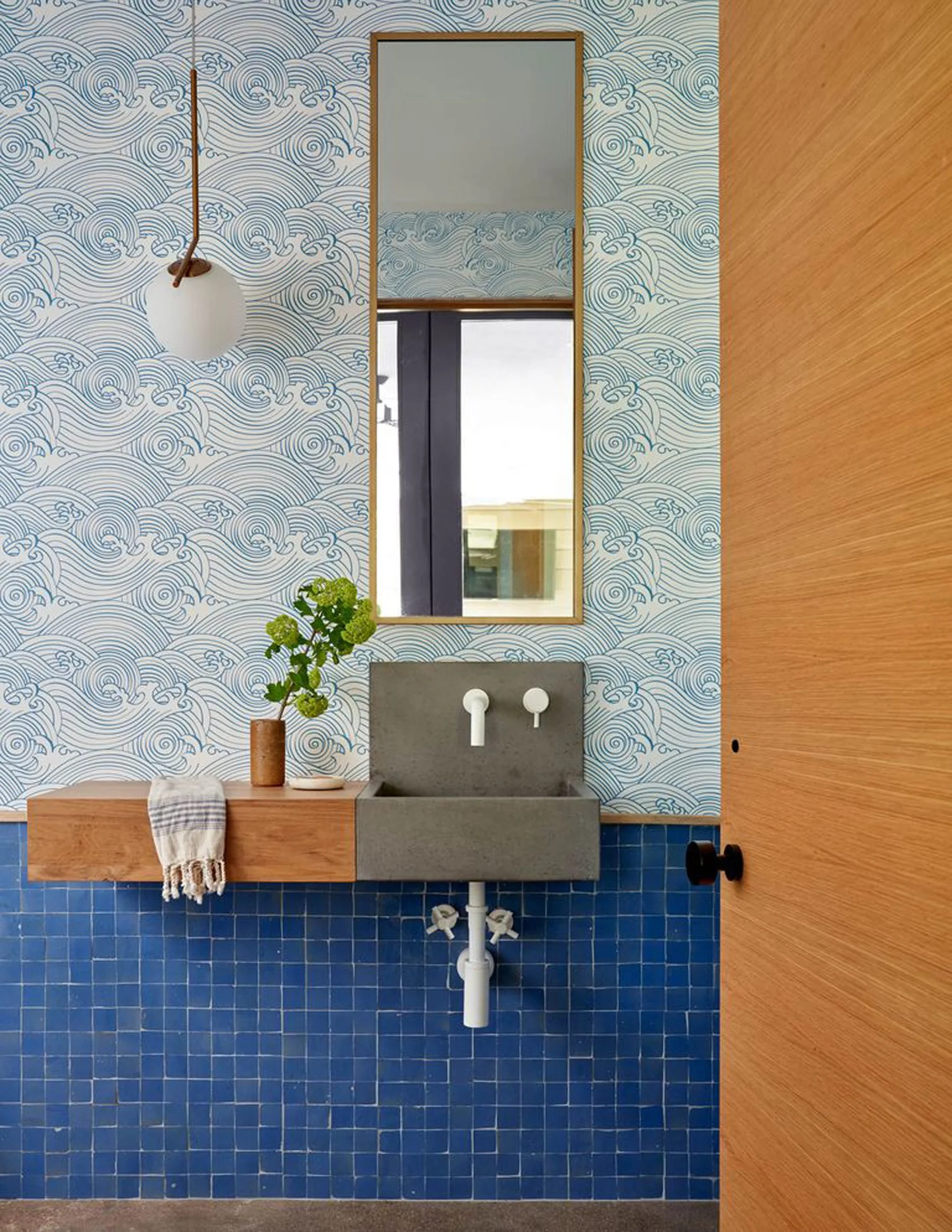A compact powder room features a grey concrete sink next to a floating wood shelf set against blue square tiles and wave-patterned wallpaper.