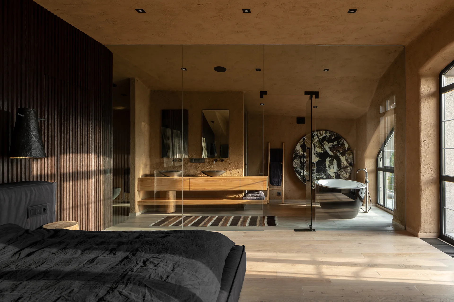 A modern bedroom with a dark fluted accent wall next to a glass-partitioned bathroom featuring textured plaster walls and a black tub.