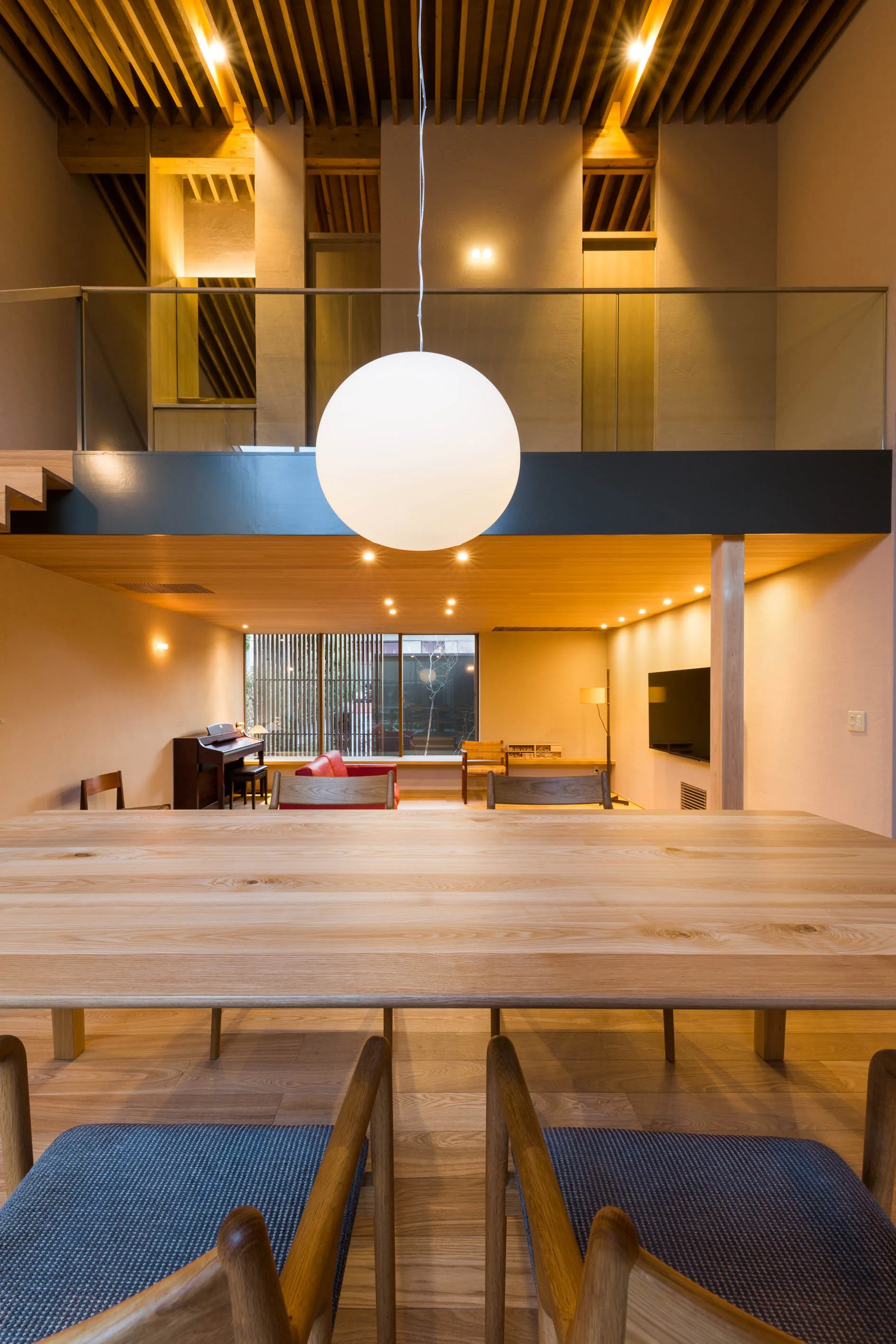 A large wooden dining table with a round white pendant light sits in an open-concept interior featuring a mezzanine level and wooden slatted ceiling.