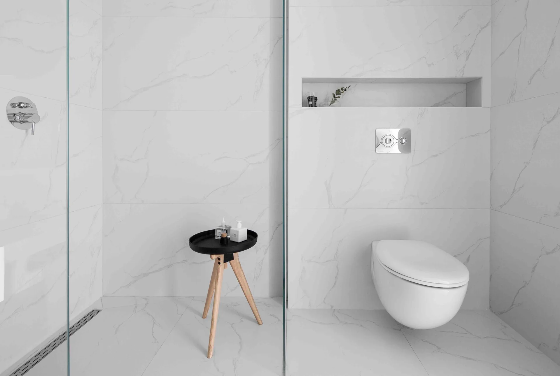 A modern bathroom features white marble-veined wall tiles, a wall-mounted toilet, a glass shower divider, and a small tripod side table.