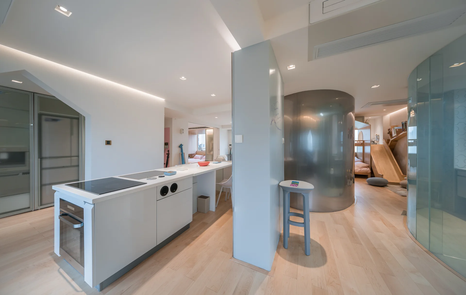 A modern open-concept interior featuring a white kitchen island, a curved stainless steel partition, and a wooden slide in a kids' play area.