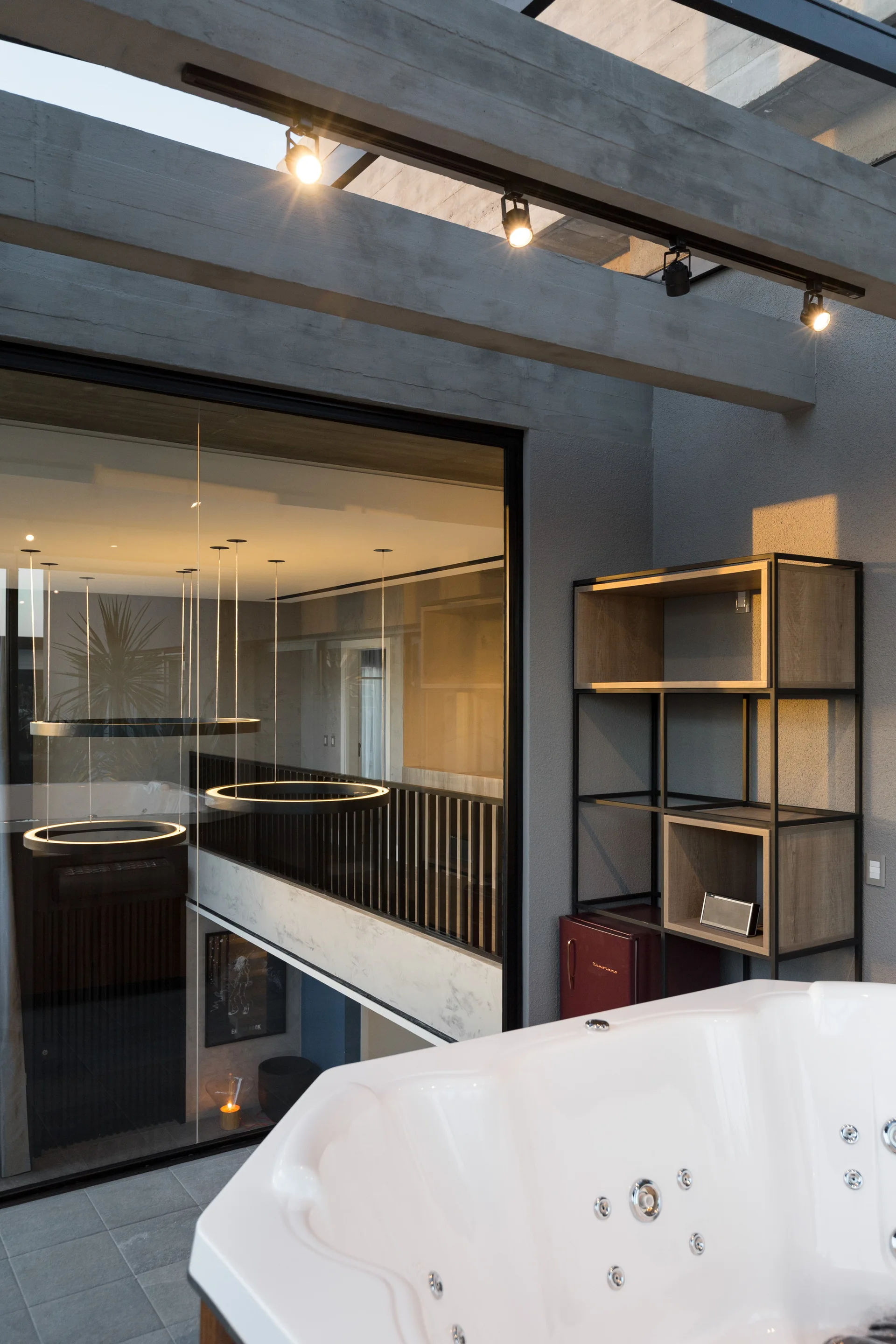 A white jetted bathtub sits next to a large glass window looking down into a living area with ring pendant lights and exposed concrete ceiling beams.