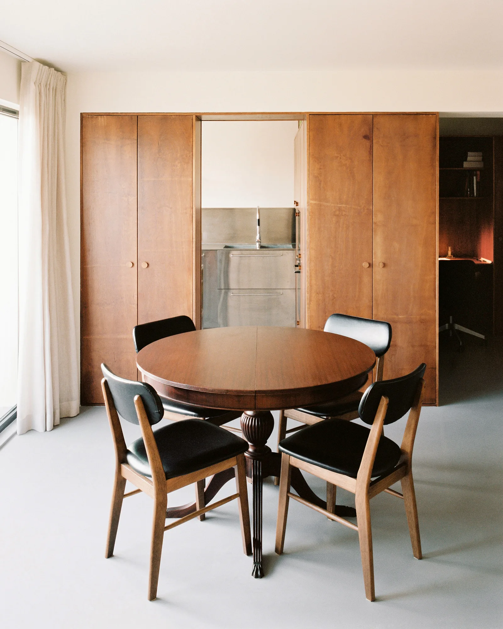 A circular wooden dining table and four black-cushioned chairs sit before a large wood storage partition wall that opens to a kitchen.