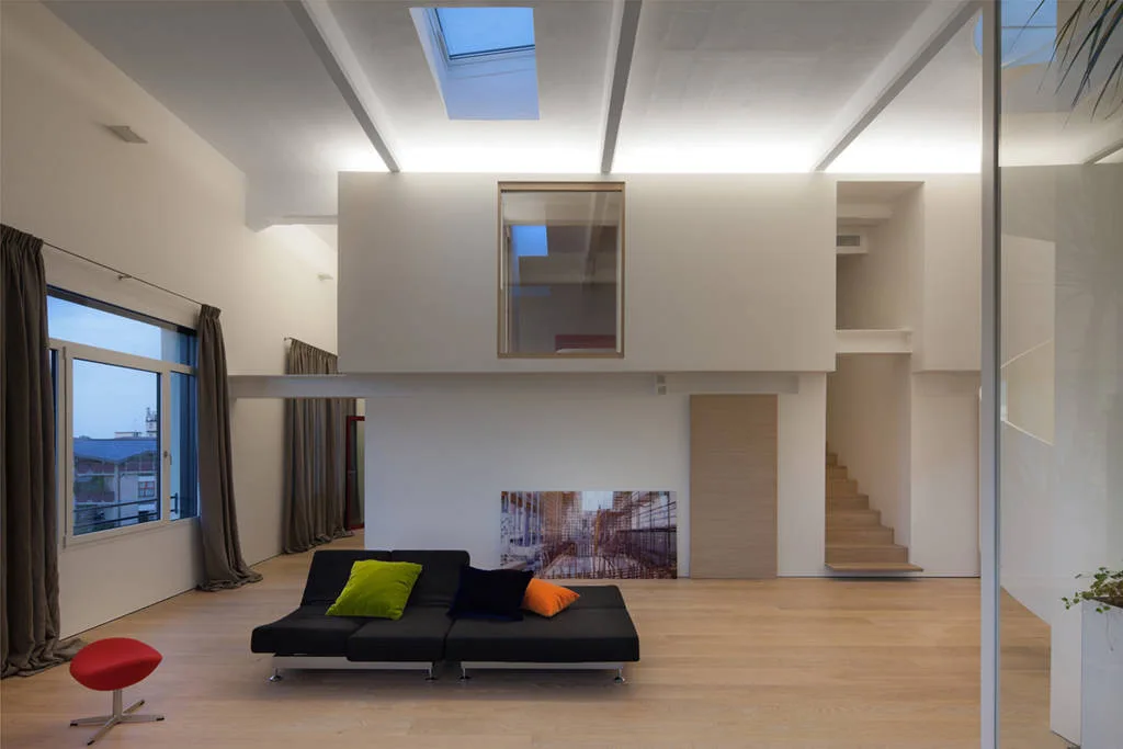 A minimalist loft living room features a low-profile black sofa, light wood floors, and a wooden floating staircase leading to an upper level.