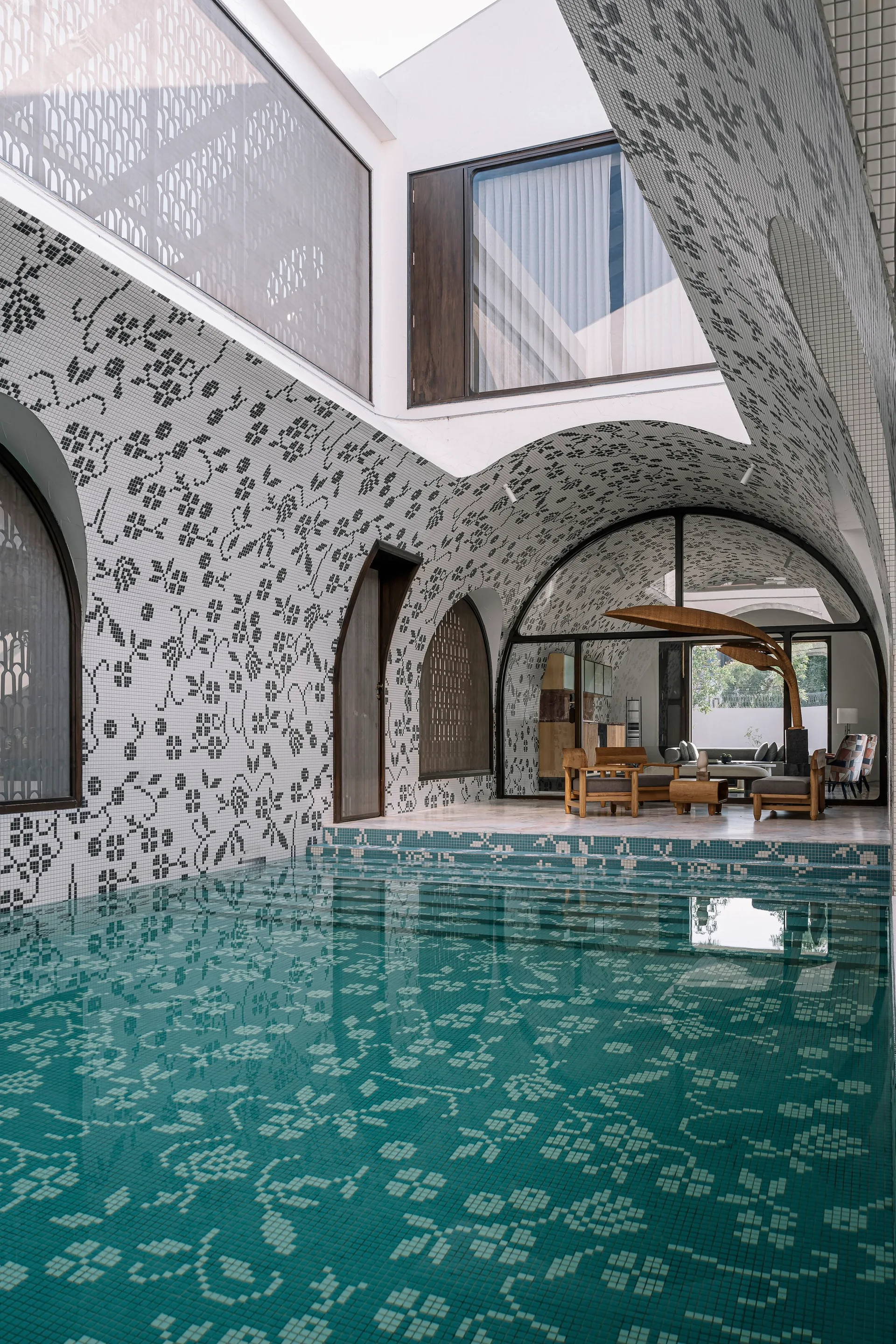 A covered outdoor swimming pool features a vaulted ceiling and walls clad in a black and white floral mosaic tile pattern next to a stone terrace.
