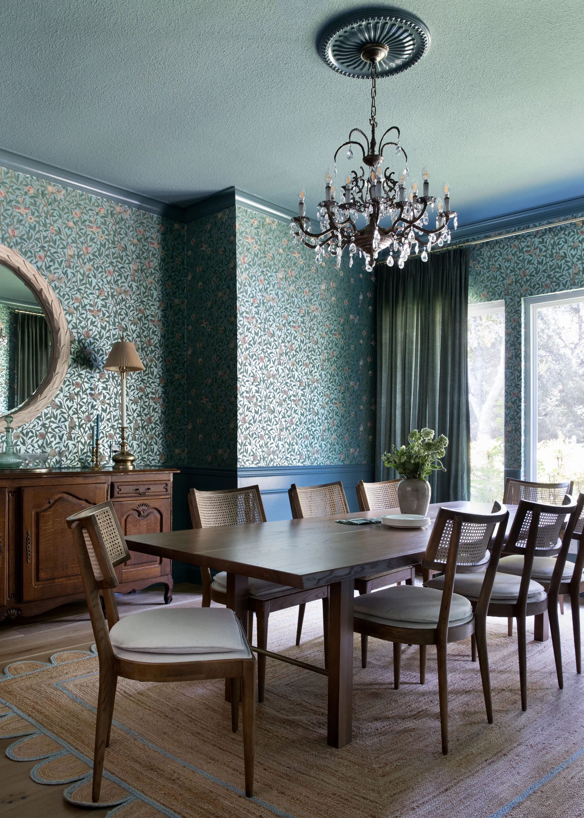 A dining room features a wooden table and cane chairs set against teal floral wallpaper and a painted teal ceiling with a chandelier.