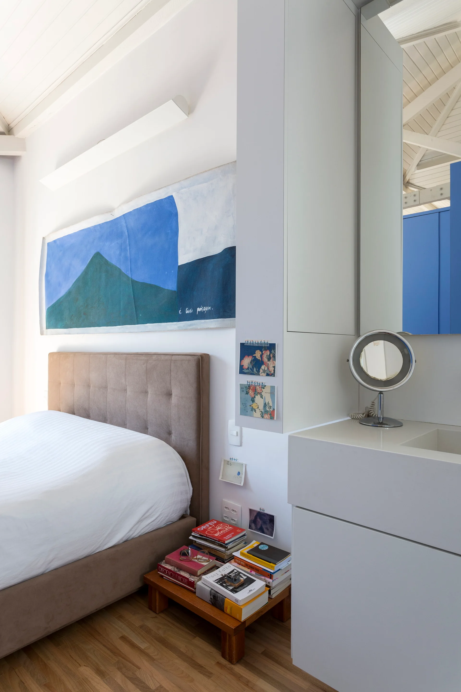 A modern minimalist bedroom features a tufted tan headboard bed, a large blue wall painting, and a white integrated vanity unit next to the bed.