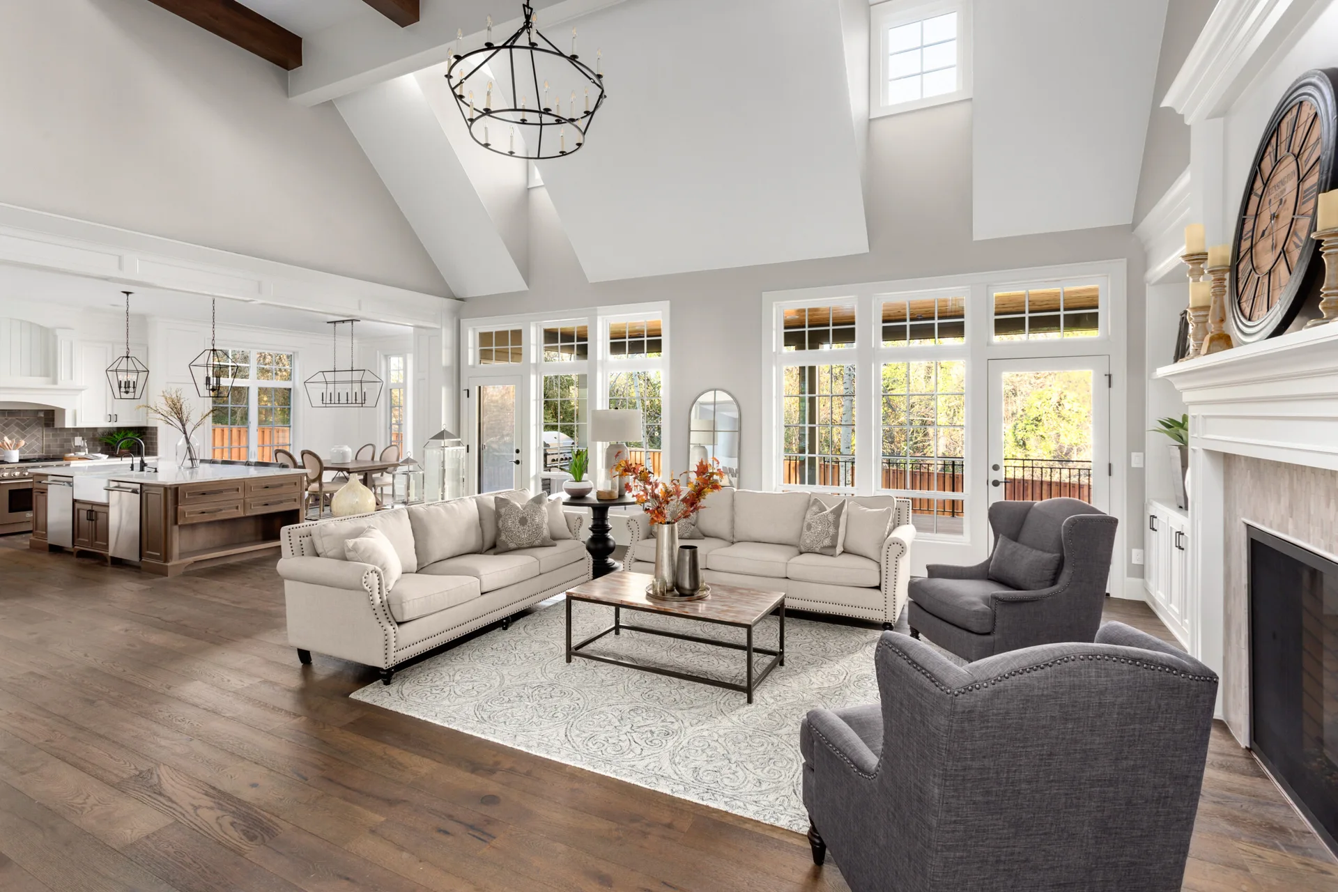 A spacious living room features two beige sofas, gray armchairs, a fireplace with a large clock, and a vaulted ceiling with dark wood beams.