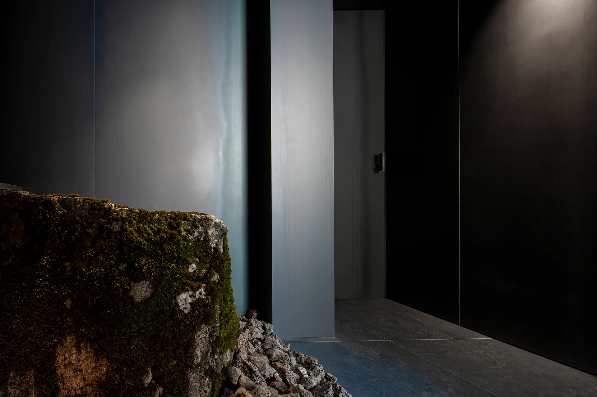 A large mossy boulder sits on small stones in a minimalist entryway next to smooth, dark charcoal matte wall panels and a recessed door.