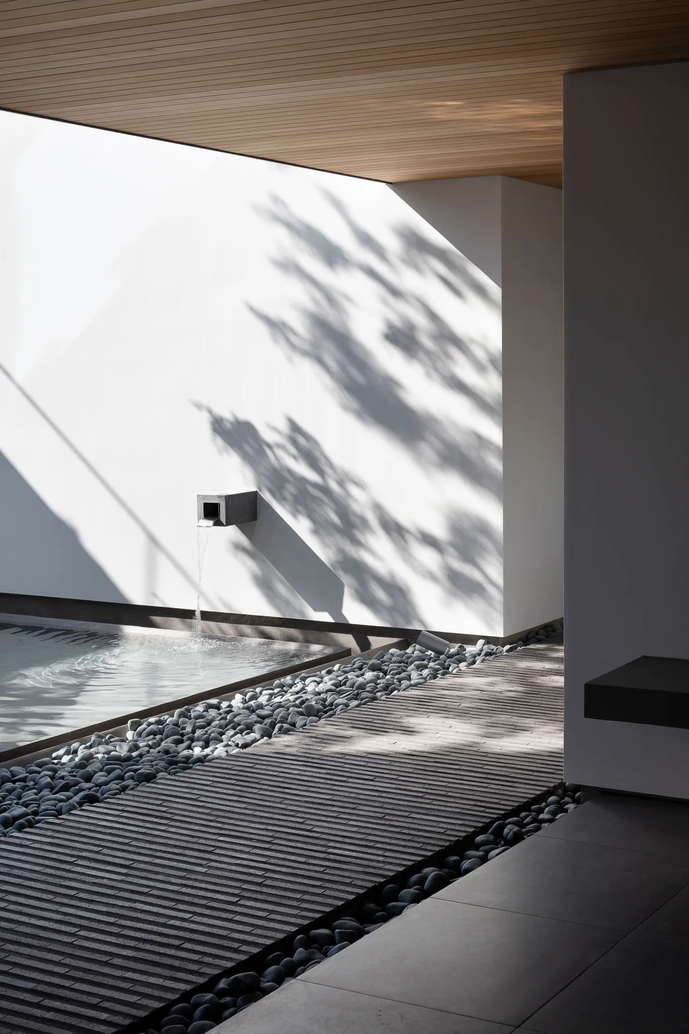 A modern outdoor courtyard with a white wall casting tree shadows, a minimalist water spout pouring into a pool, and a brick walkway flanked by pebbles.