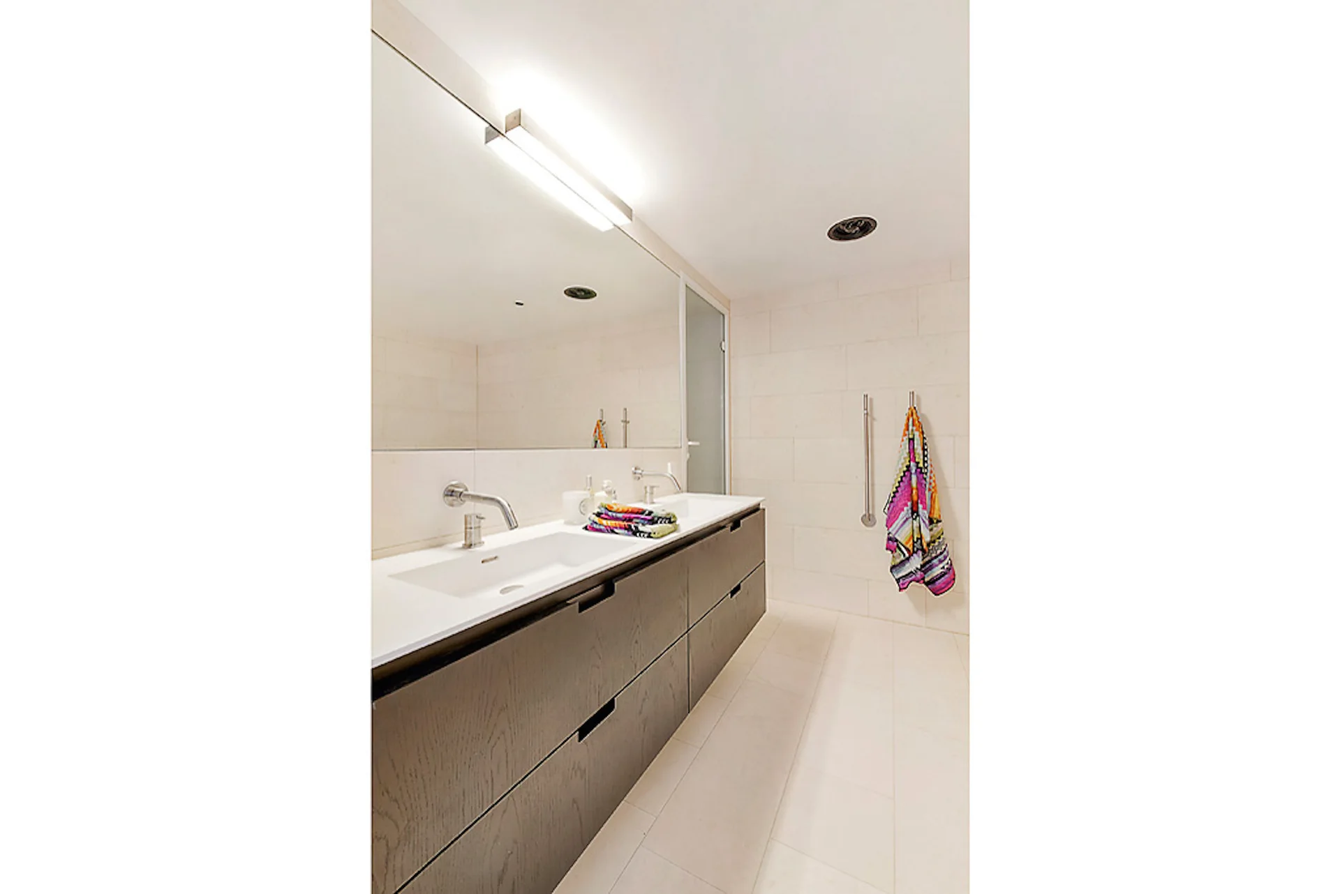 A contemporary bathroom featuring a dark wood double vanity, large mirror, and rectangular beige wall tiles.
