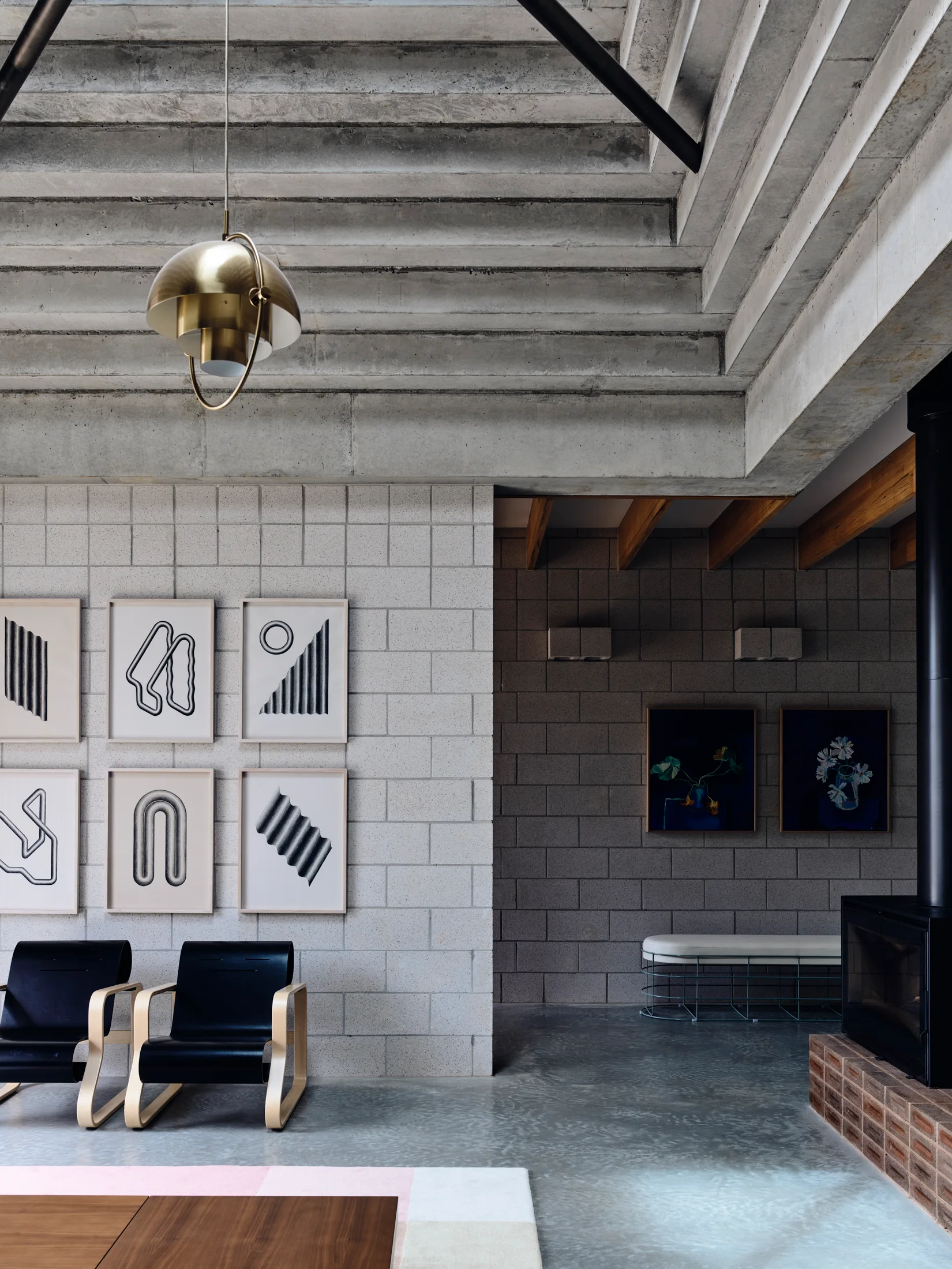 A modern living room features raw concrete block walls, a ribbed concrete ceiling, a brass pendant light, and a grid of black and white graphic art.