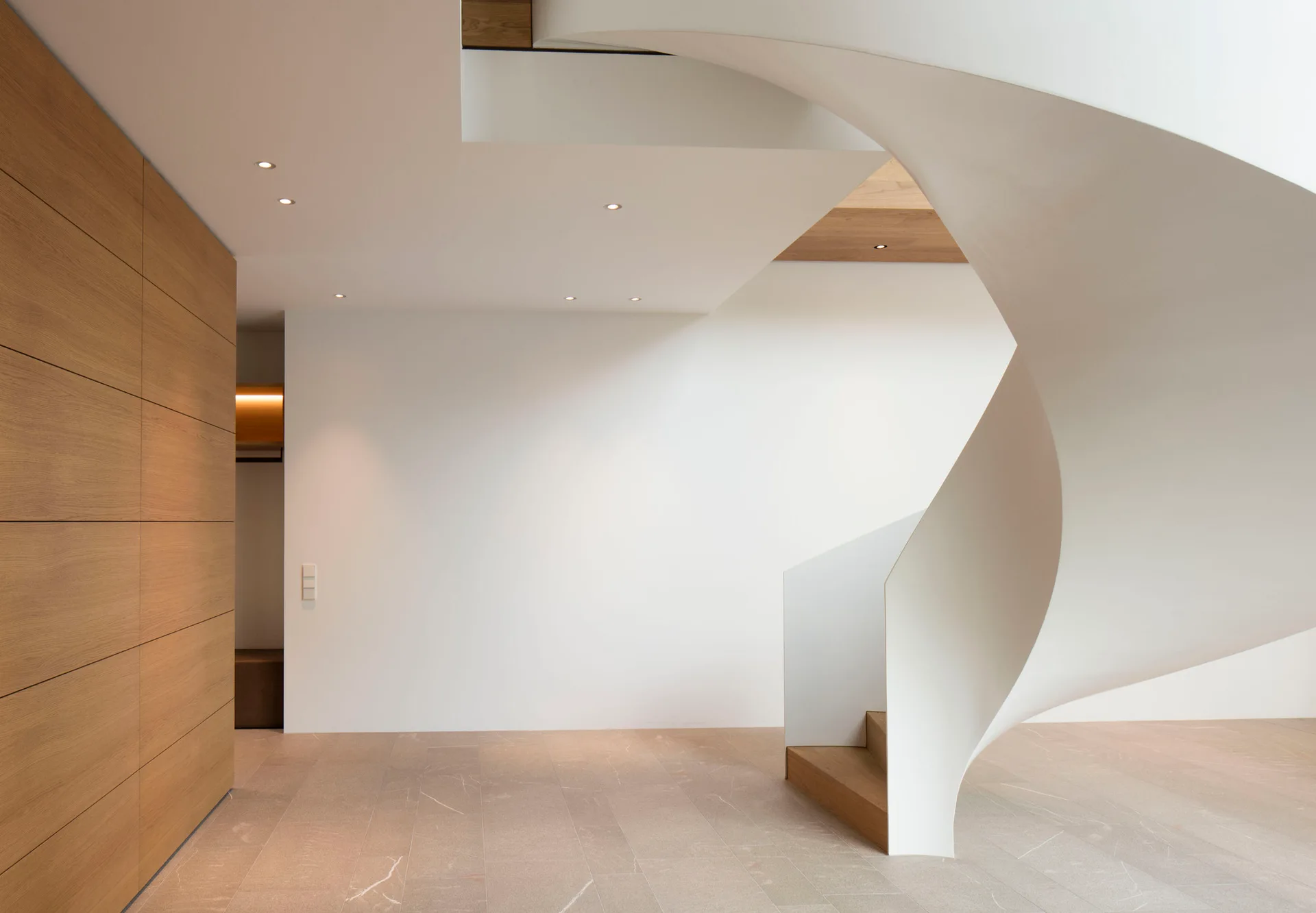 A minimalist entryway featuring a curved white sculptural spiral staircase, light wood wall paneling, and large-format beige stone floor tiles.