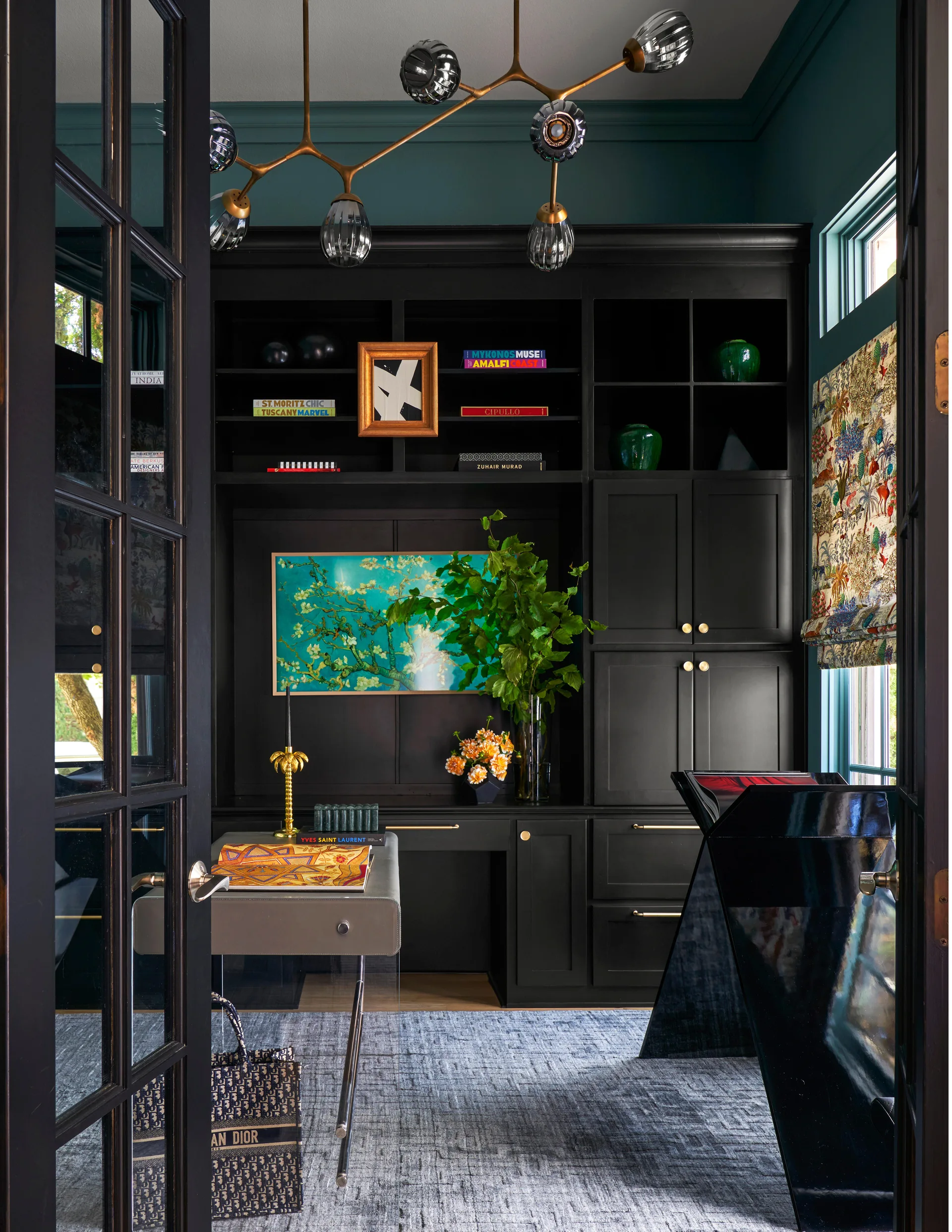 A moody home office viewed through dark French doors, featuring custom black cabinetry, a desk with glass legs, and a gold branching chandelier.