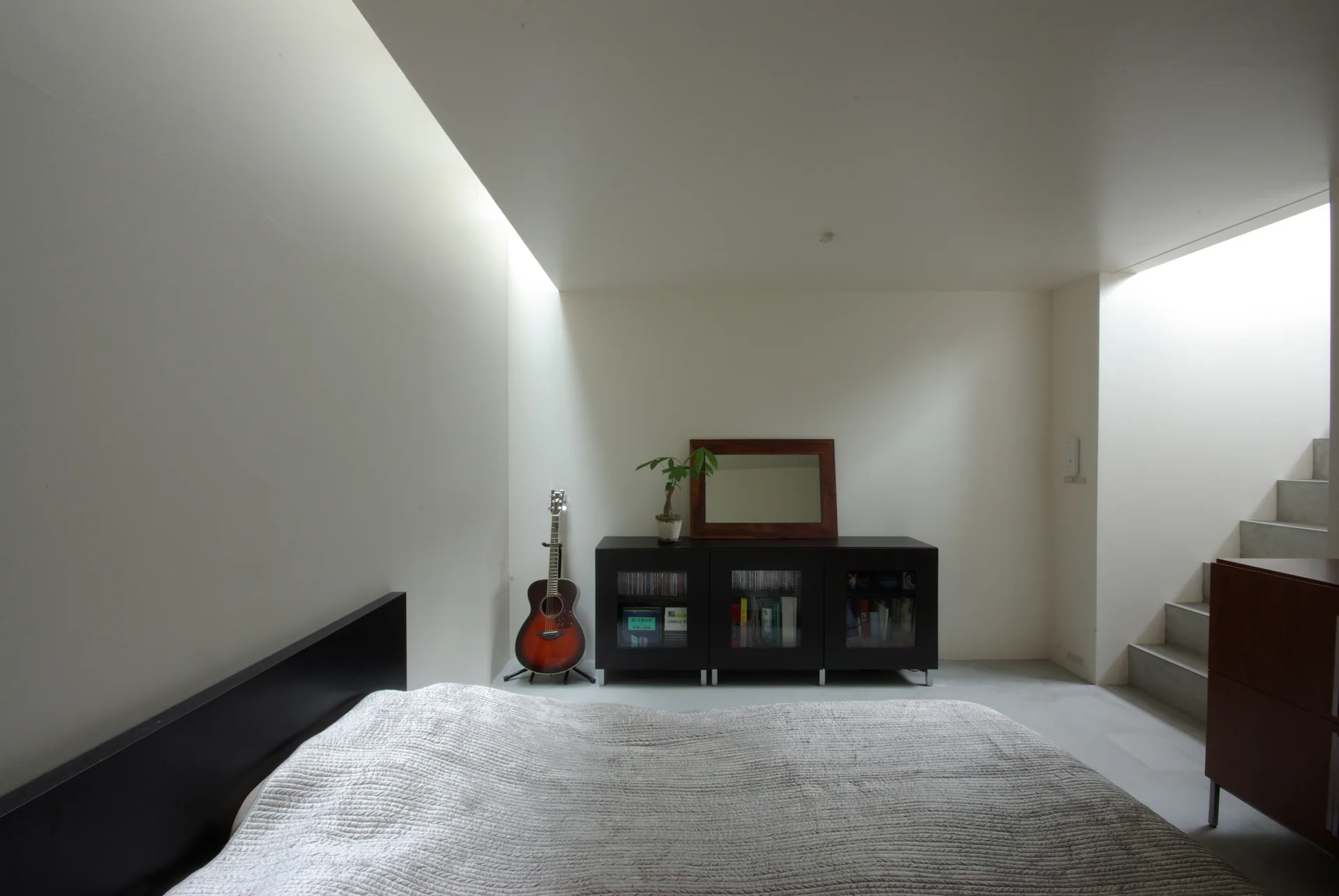 A minimalist bedroom with white walls, a textured grey bed, a black glass-front cabinet, and natural light streaming from a ceiling skylight.