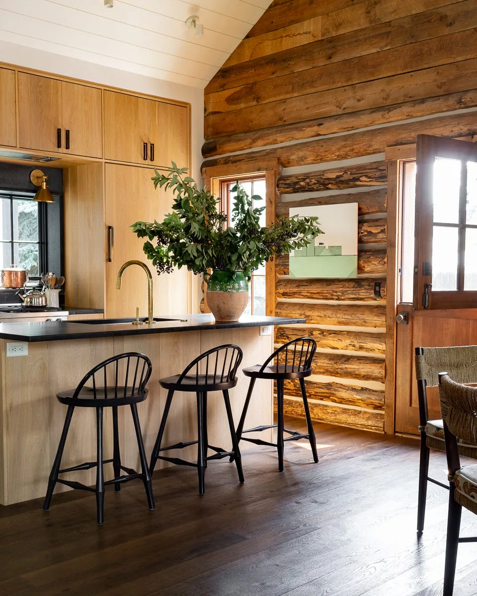A rustic kitchen features a wood island with three black spindle-back stools, set against an exposed log wall and a wooden Dutch door.