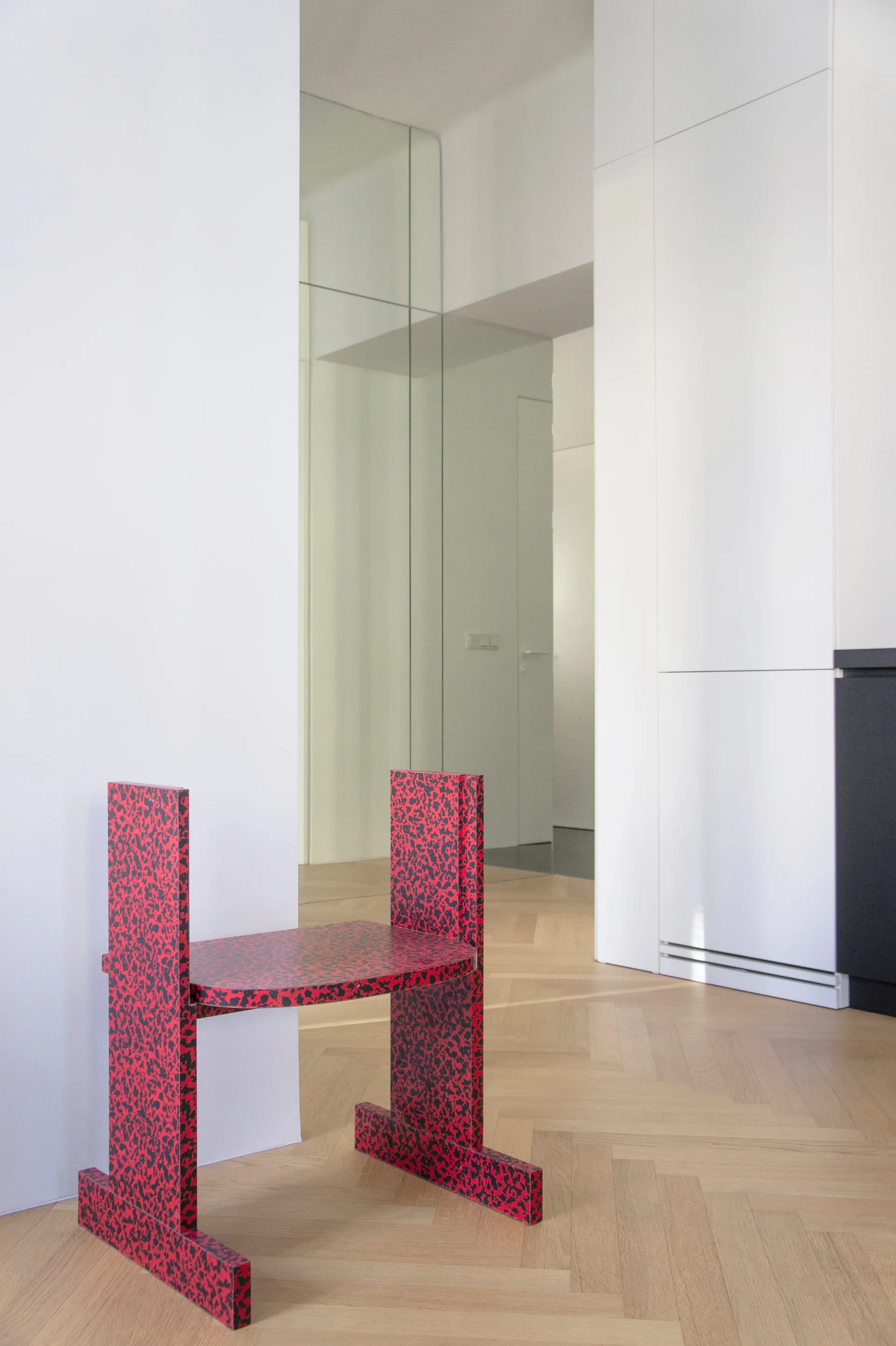 A sculptural red and black speckled chair sits on light oak herringbone parquet flooring in a minimalist white hallway with built-in cabinetry.