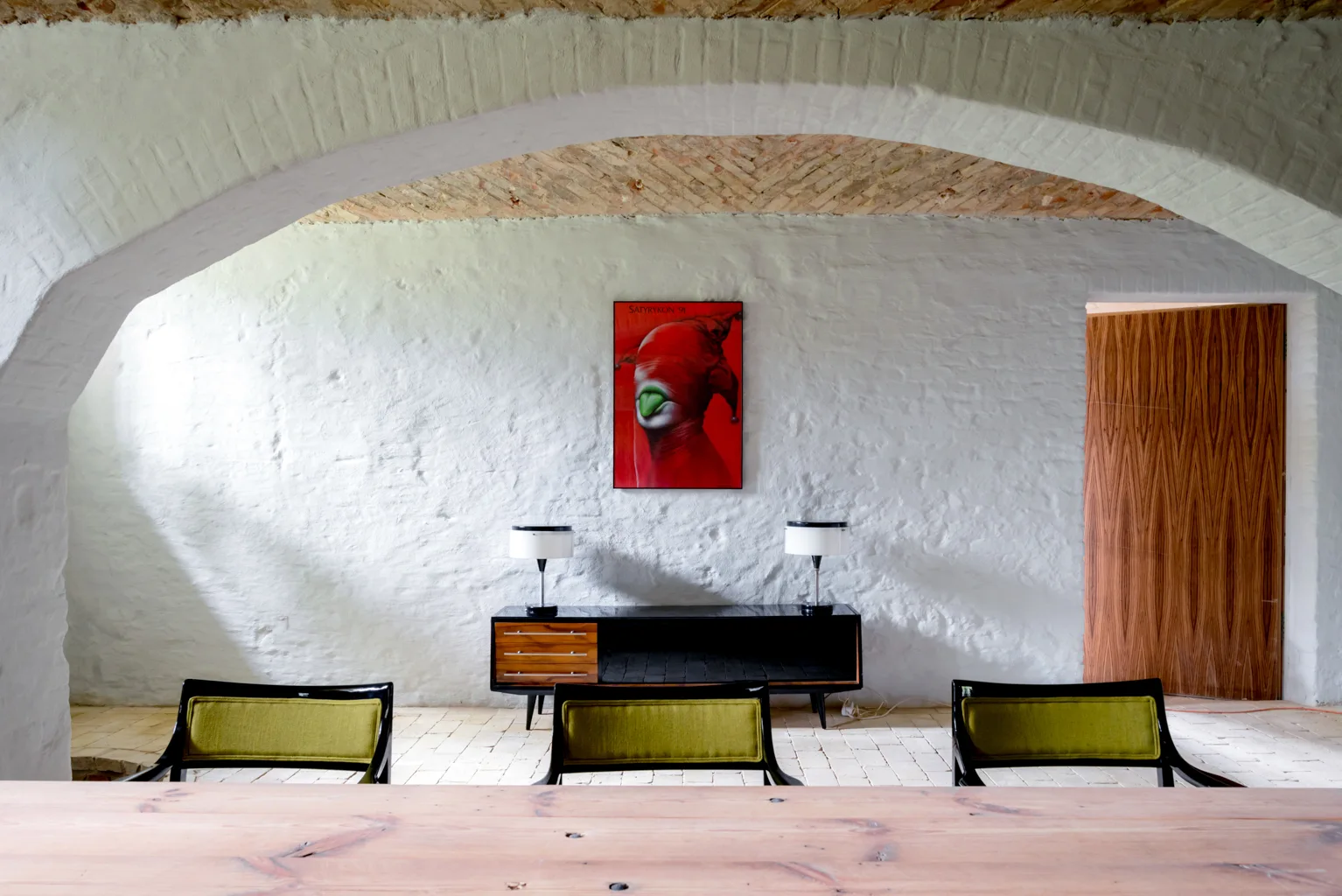 A minimalist room features a white painted brick wall, a brick vaulted ceiling, a black sideboard with red art, and olive green chairs.