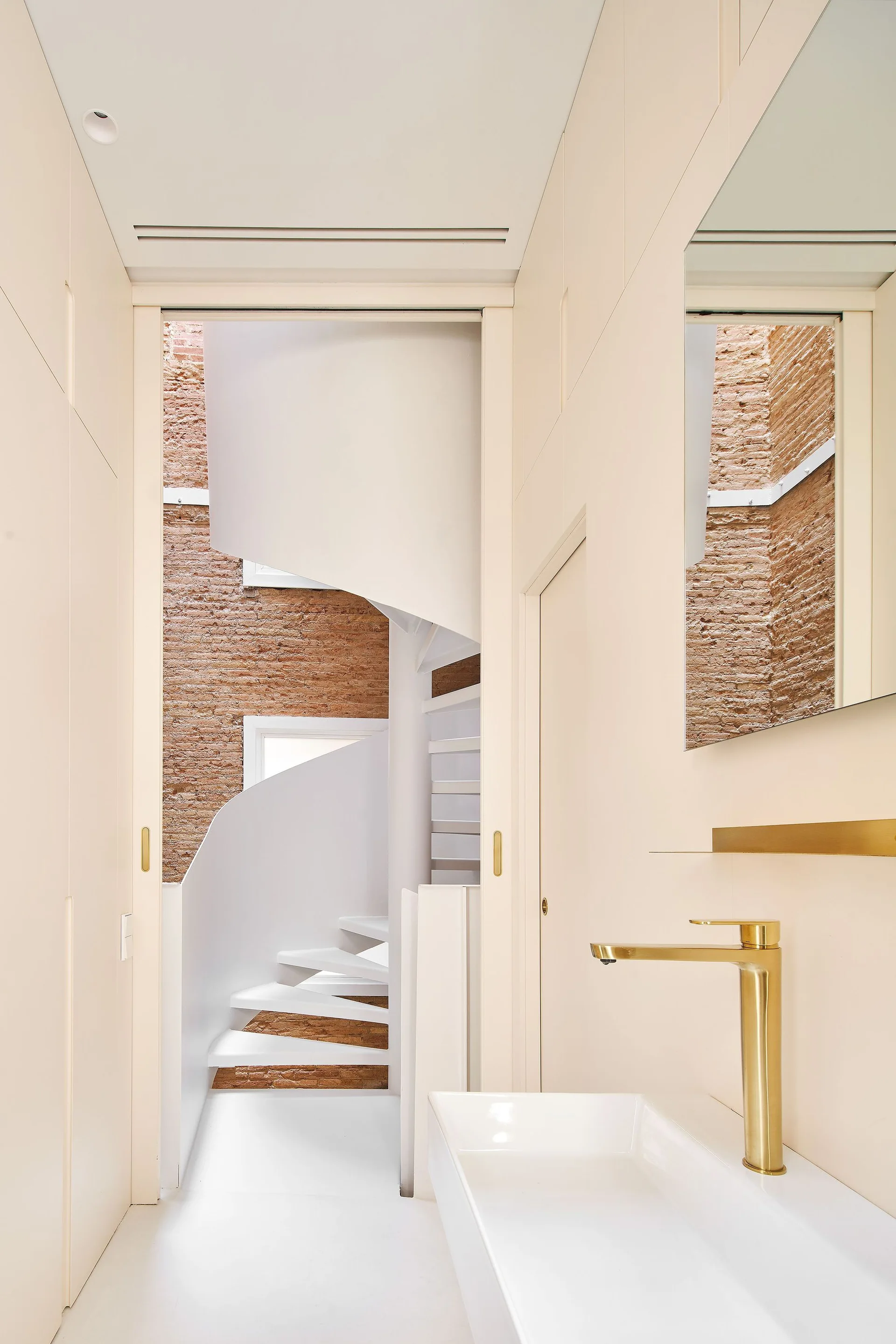 A minimalist beige powder room with a brass faucet and mirror opens to a white spiral staircase set against a rustic exposed brick wall.