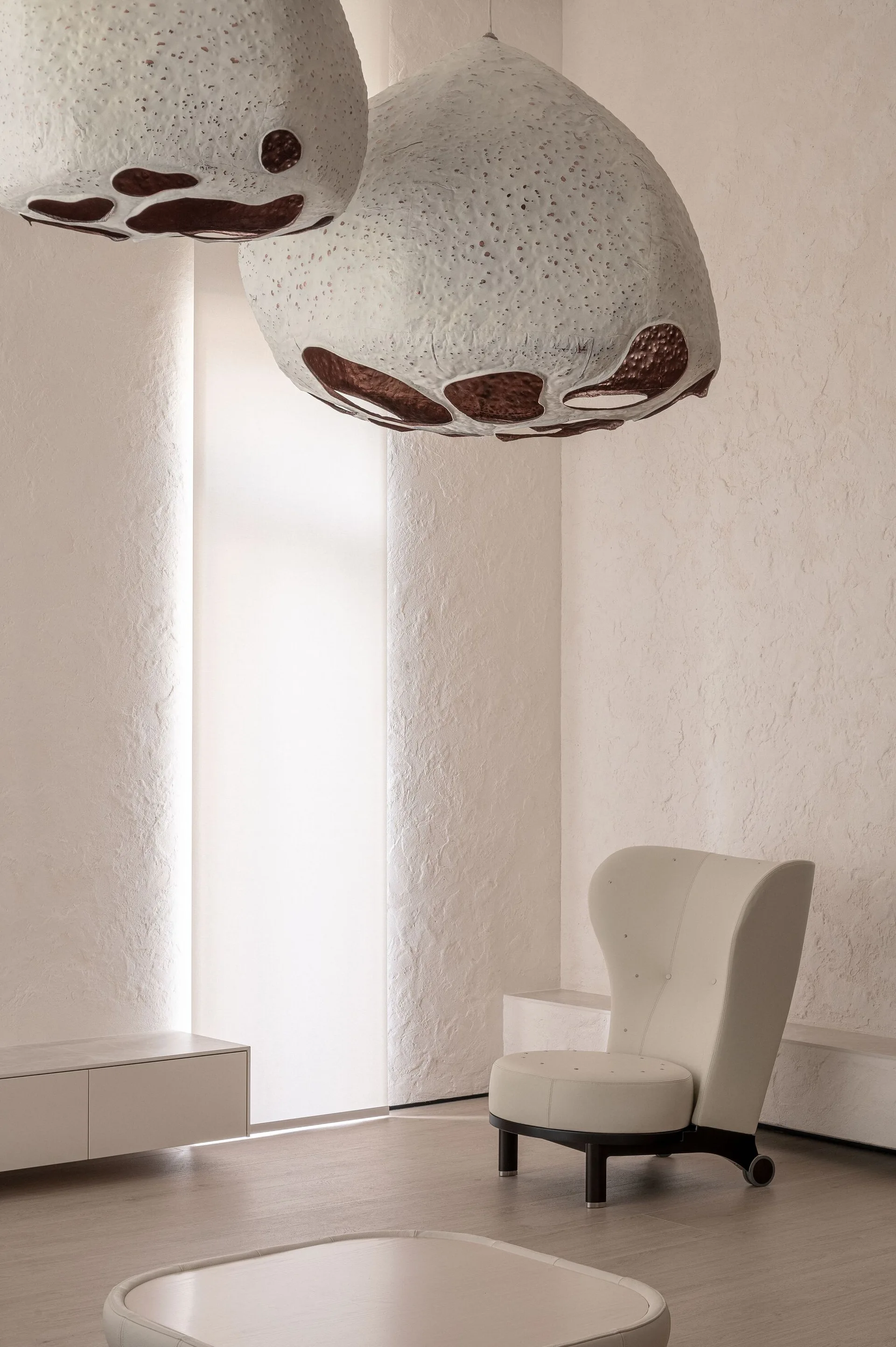 A minimalist off-white room features large bulbous pendant lights, a cream wingback chair, and textured plaster walls with a glowing light alcove.