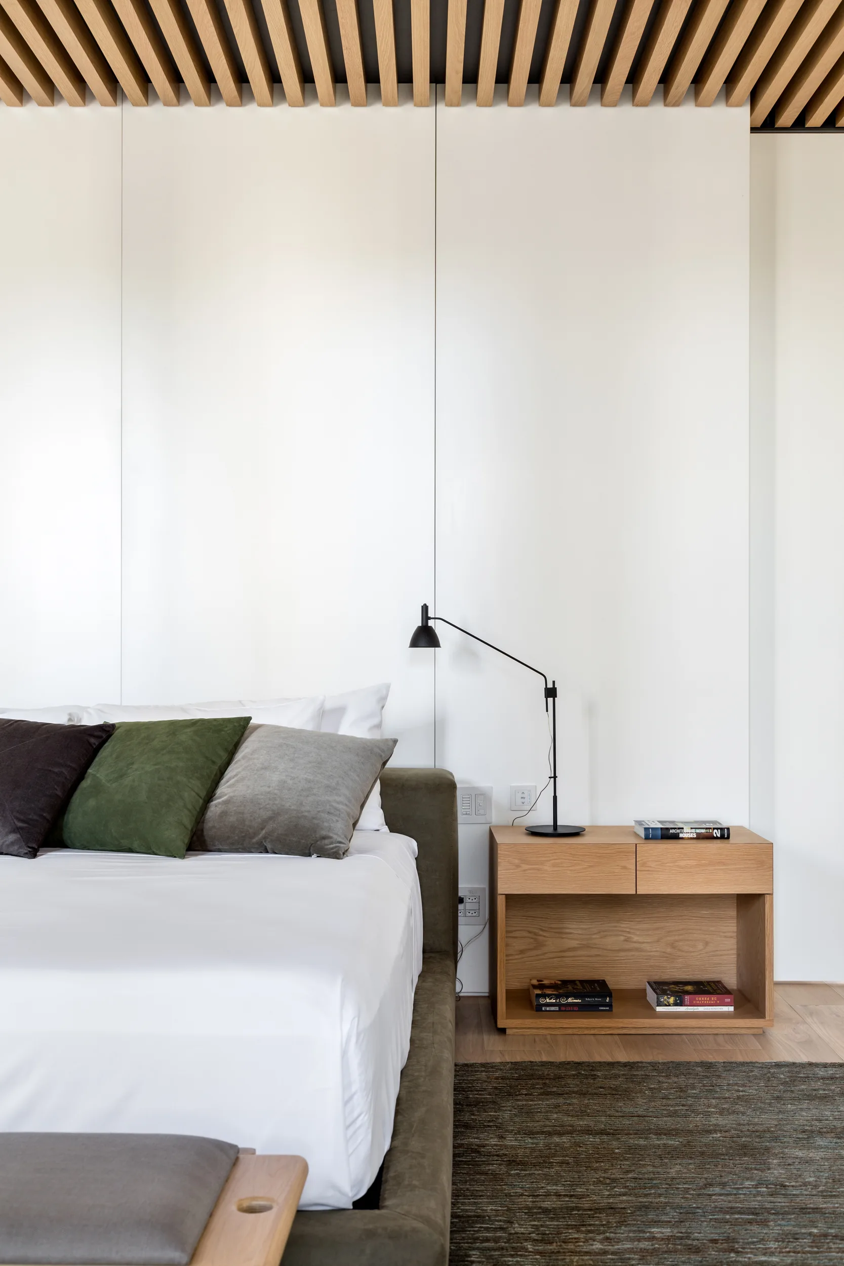 A modern bedroom features a low-profile upholstered bed, a light wood nightstand with a black lamp, and a linear slatted wood ceiling.