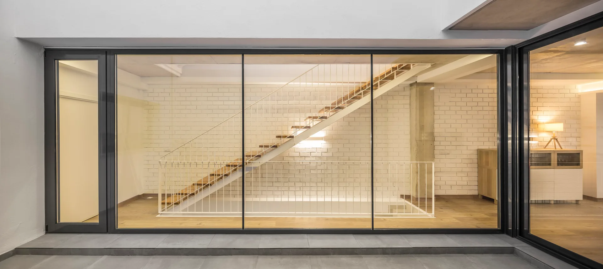 A modern interior staircase with wooden treads and a white metal railing is visible through a large, floor-to-ceiling glass window partition.