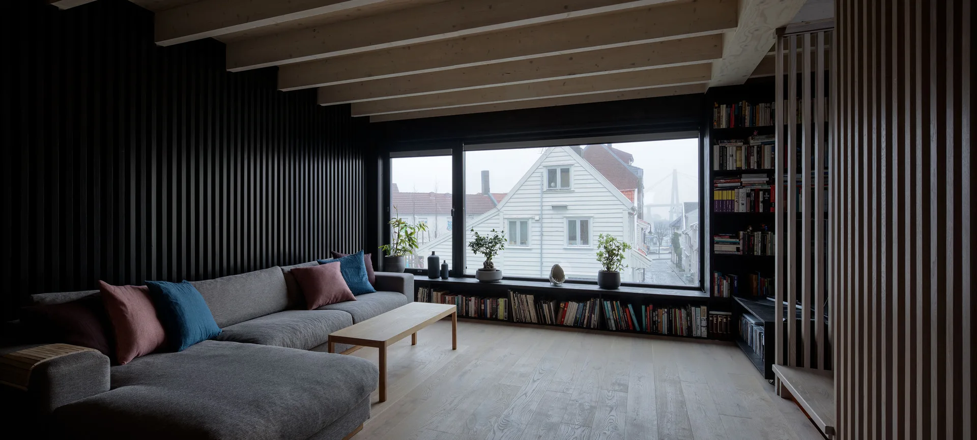 A minimalist living room with a grey sectional, black slatted wall paneling, exposed wooden ceiling beams, and a low bookshelf beneath a large window.