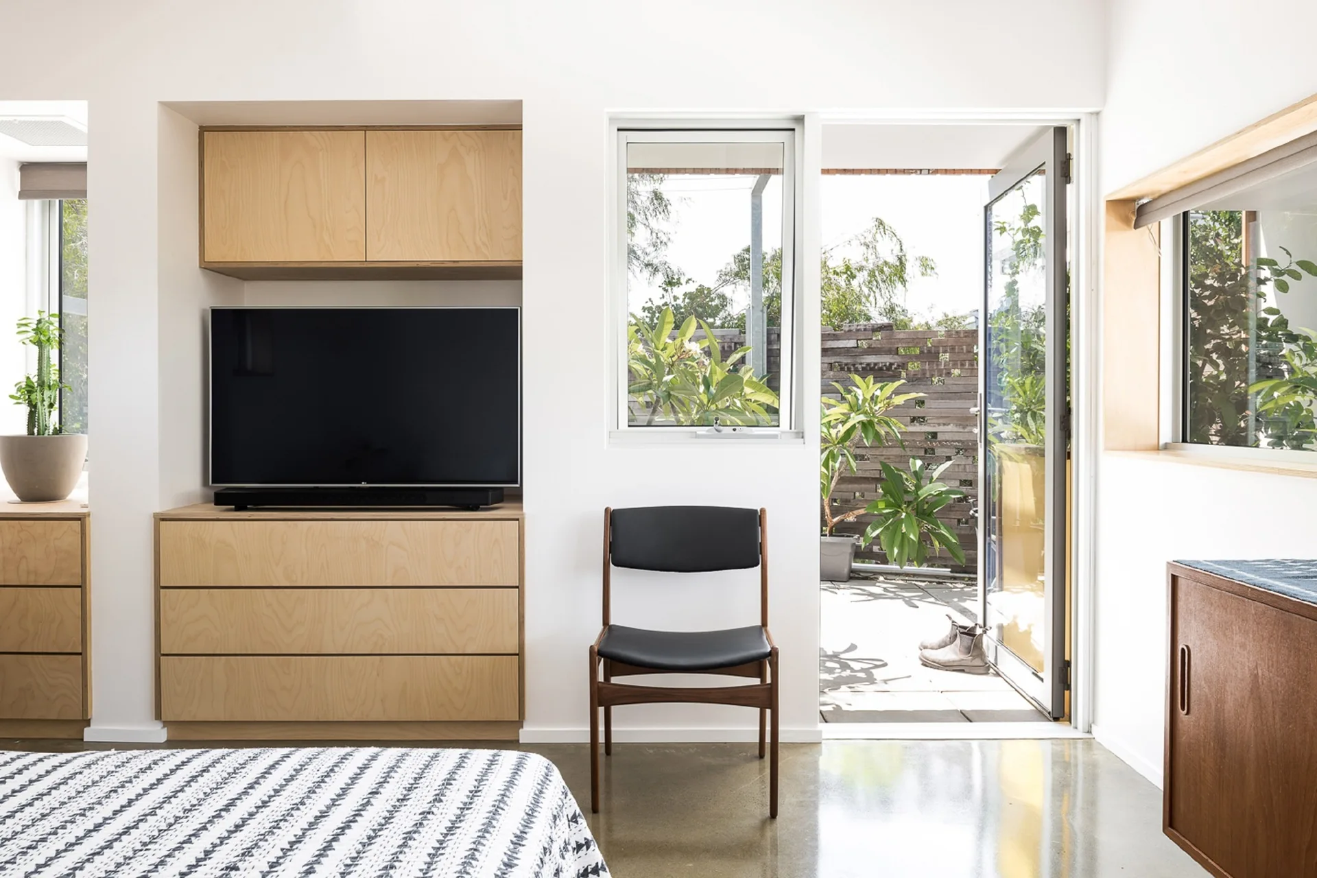A modern bedroom features custom plywood built-in cabinetry, a black mid-century chair, and an open glass door leading to a patio.