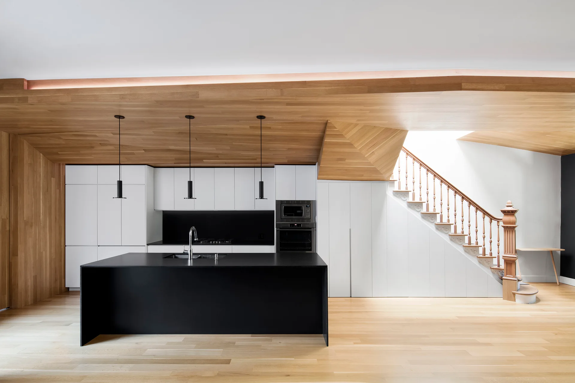 A modern kitchen featuring a black central island, integrated white cabinets, a sloped wood ceiling, and a wooden staircase illuminated by a skylight.