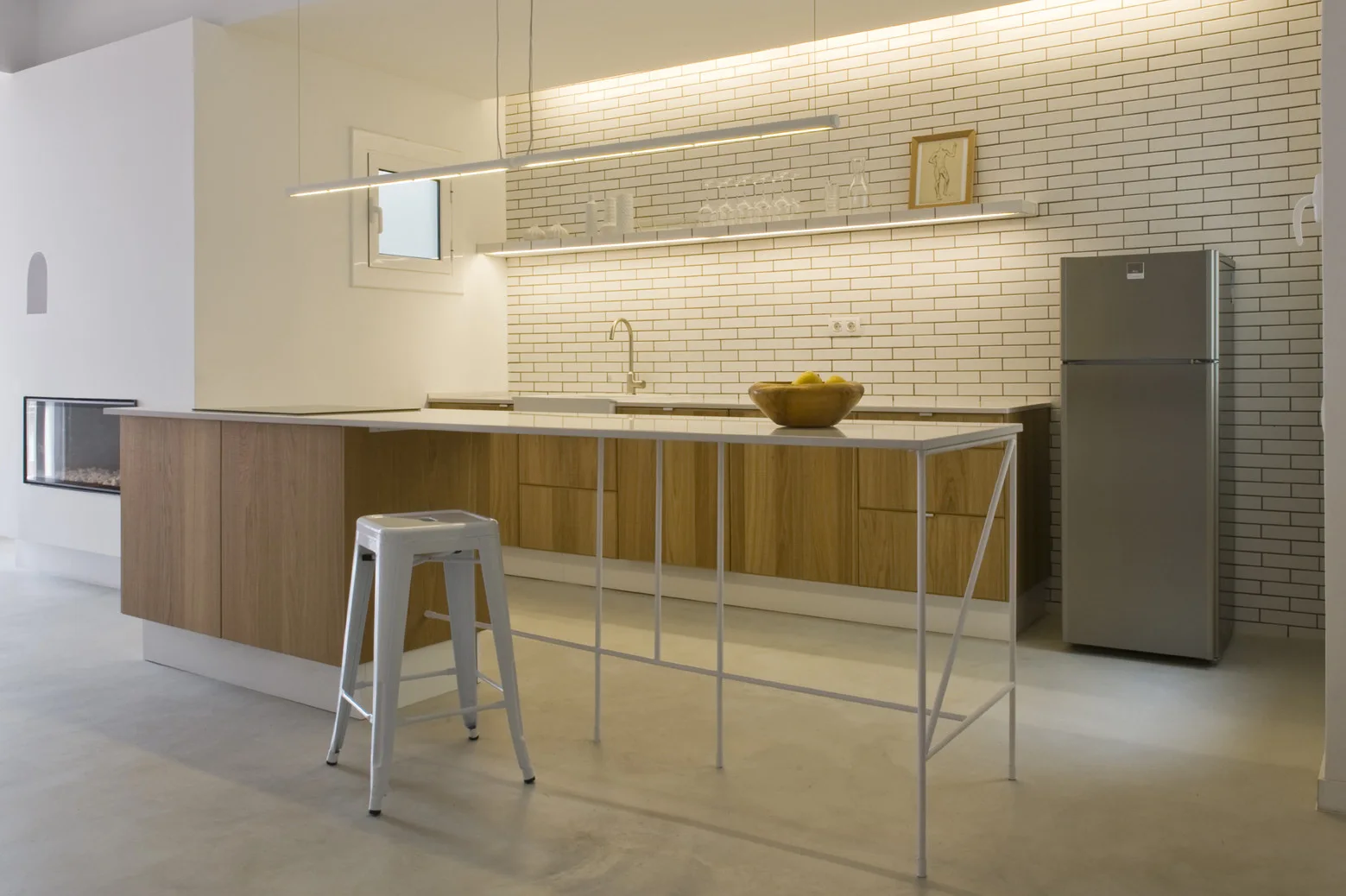 A minimalist kitchen features wood lower cabinets, a white subway tile backsplash, a floating shelf with integrated lighting, and a white metal island.