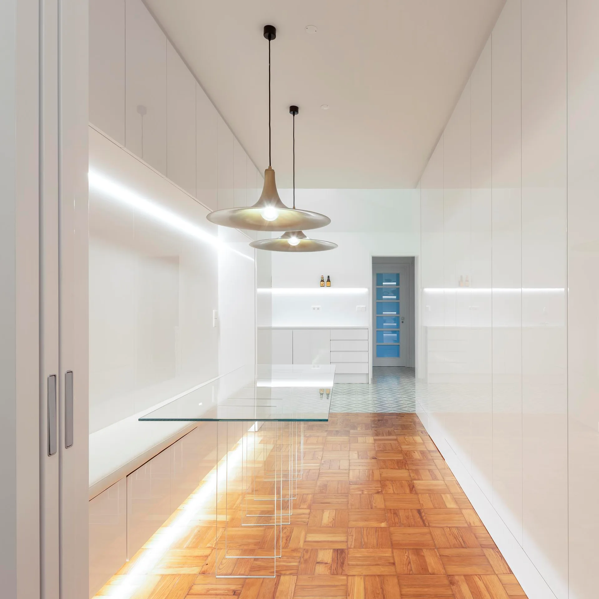 A minimalist white hallway features glossy built-in cabinetry, a long glass console table, brass pendant lights, and classic parquet wood flooring.