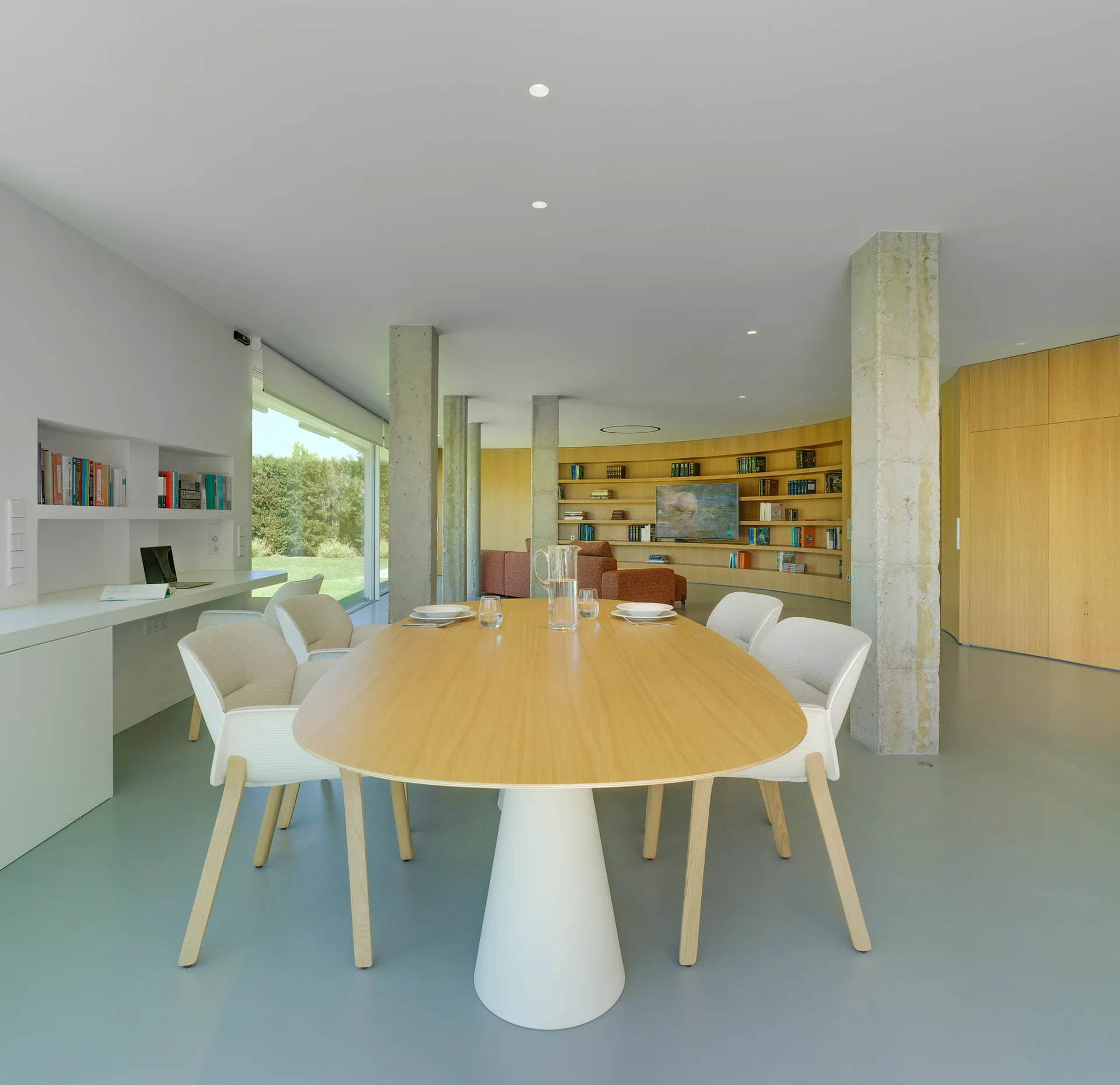 An oval light wood dining table with white chairs stands in an open-plan room flanked by raw concrete columns and a curved wood-paneled wall.