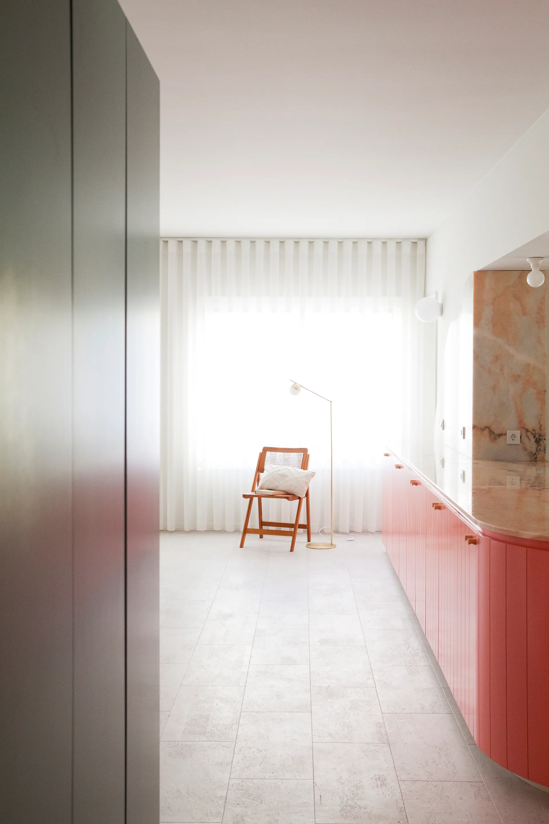 A bright minimalist kitchen featuring salmon pink lower cabinets with a matching pink-toned marble countertop, a wooden cane chair, and sheer white curtains.