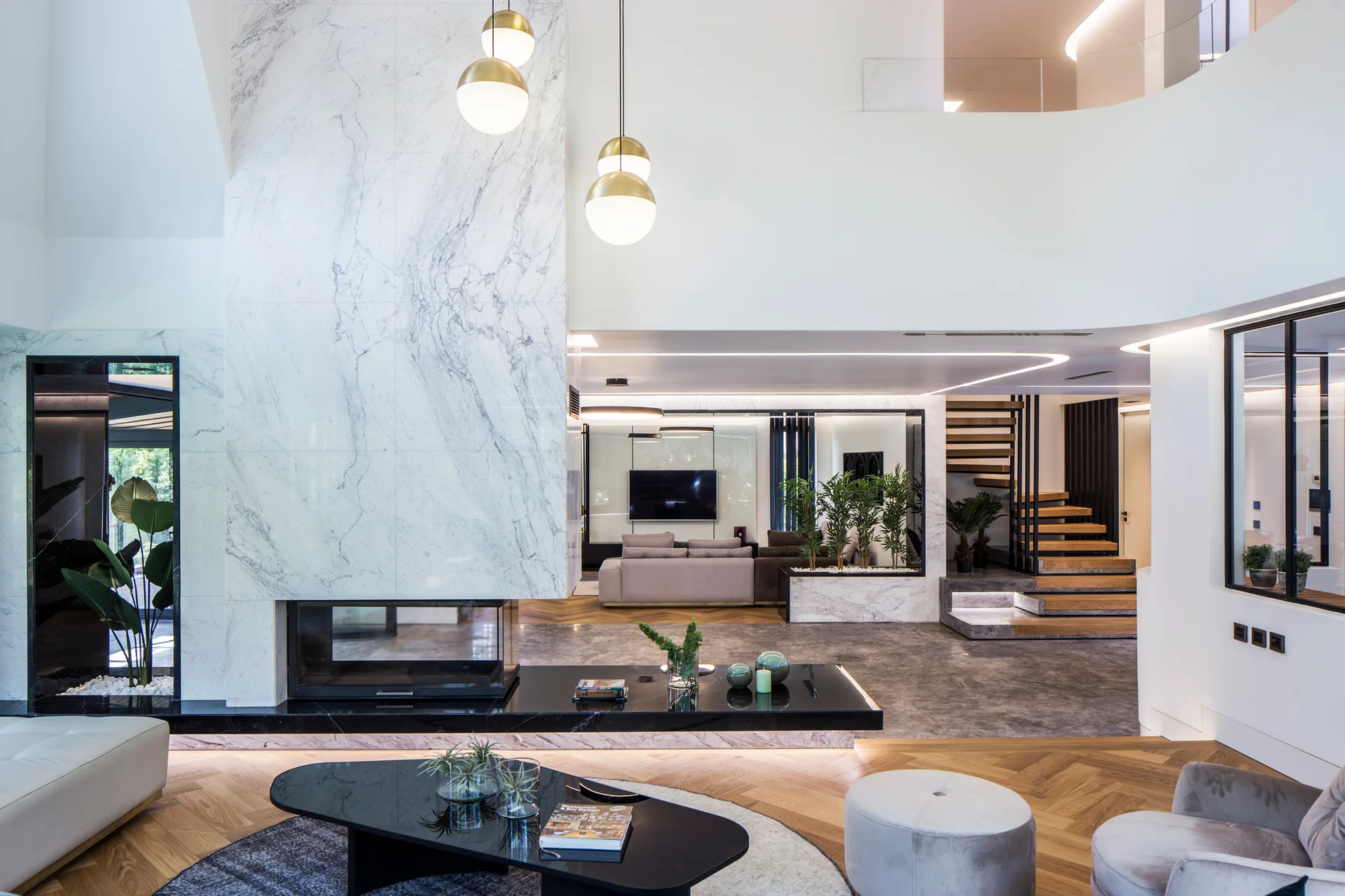 A spacious modern living room featuring a marble-clad fireplace, a floating wooden staircase, and herringbone flooring transitioning to grey stone.