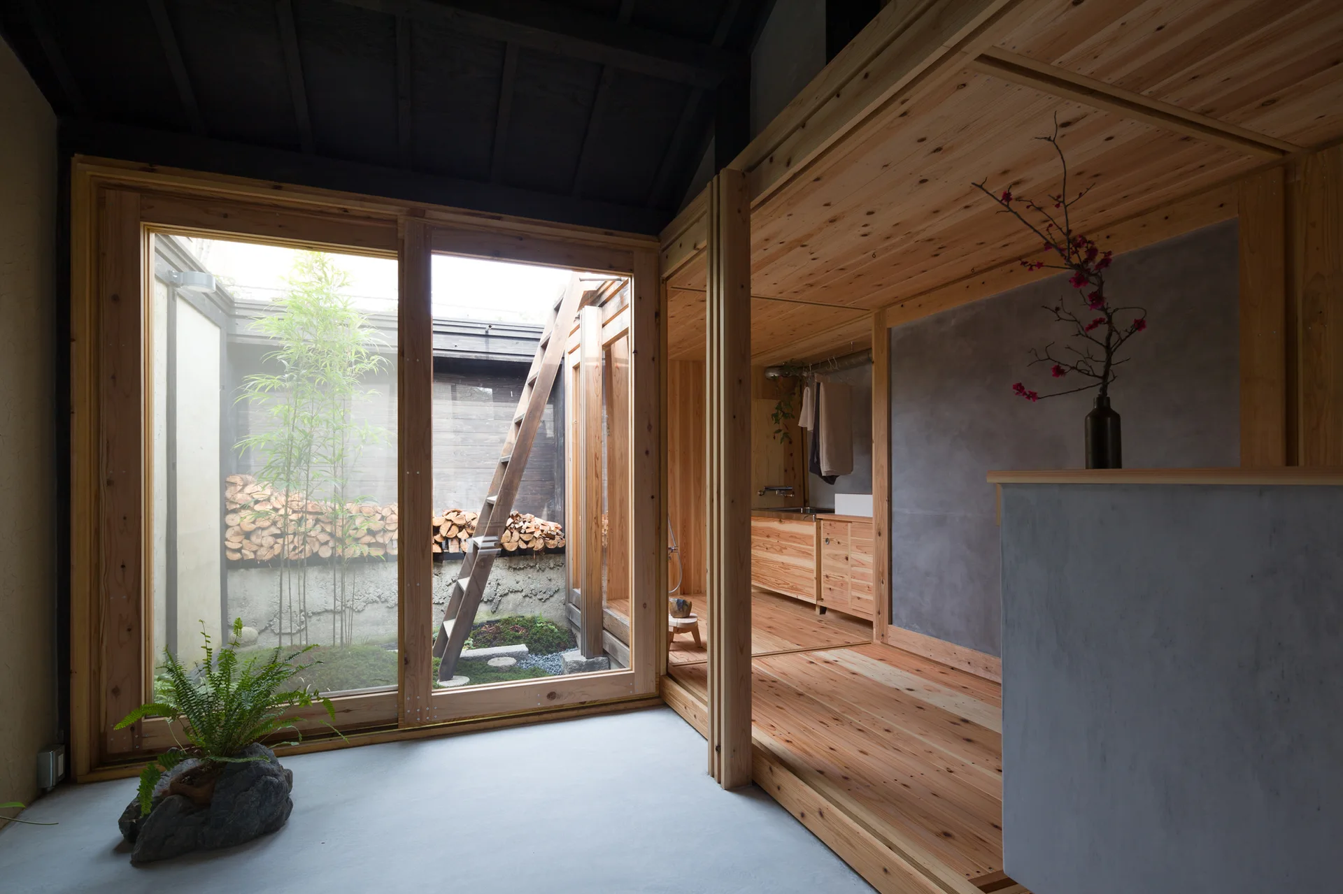A minimalist room with light wood floors and grey concrete walls features a large glass sliding door looking out onto a small courtyard with bamboo.