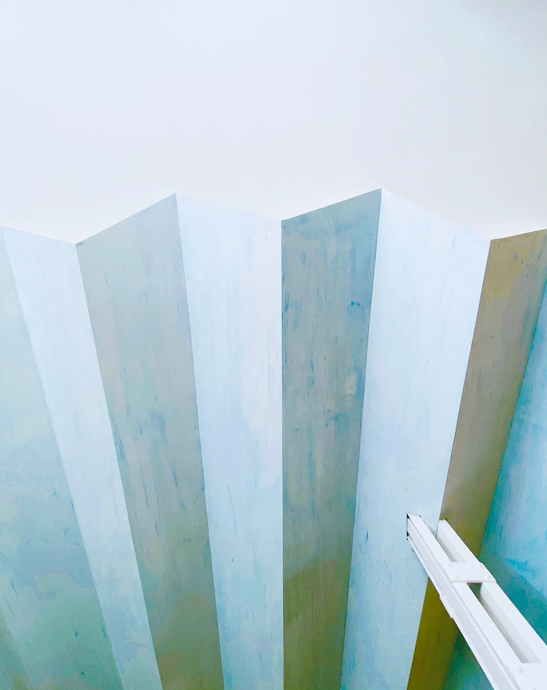 Vertical faceted wood panels with a washed blue paint finish meeting a flat white ceiling.