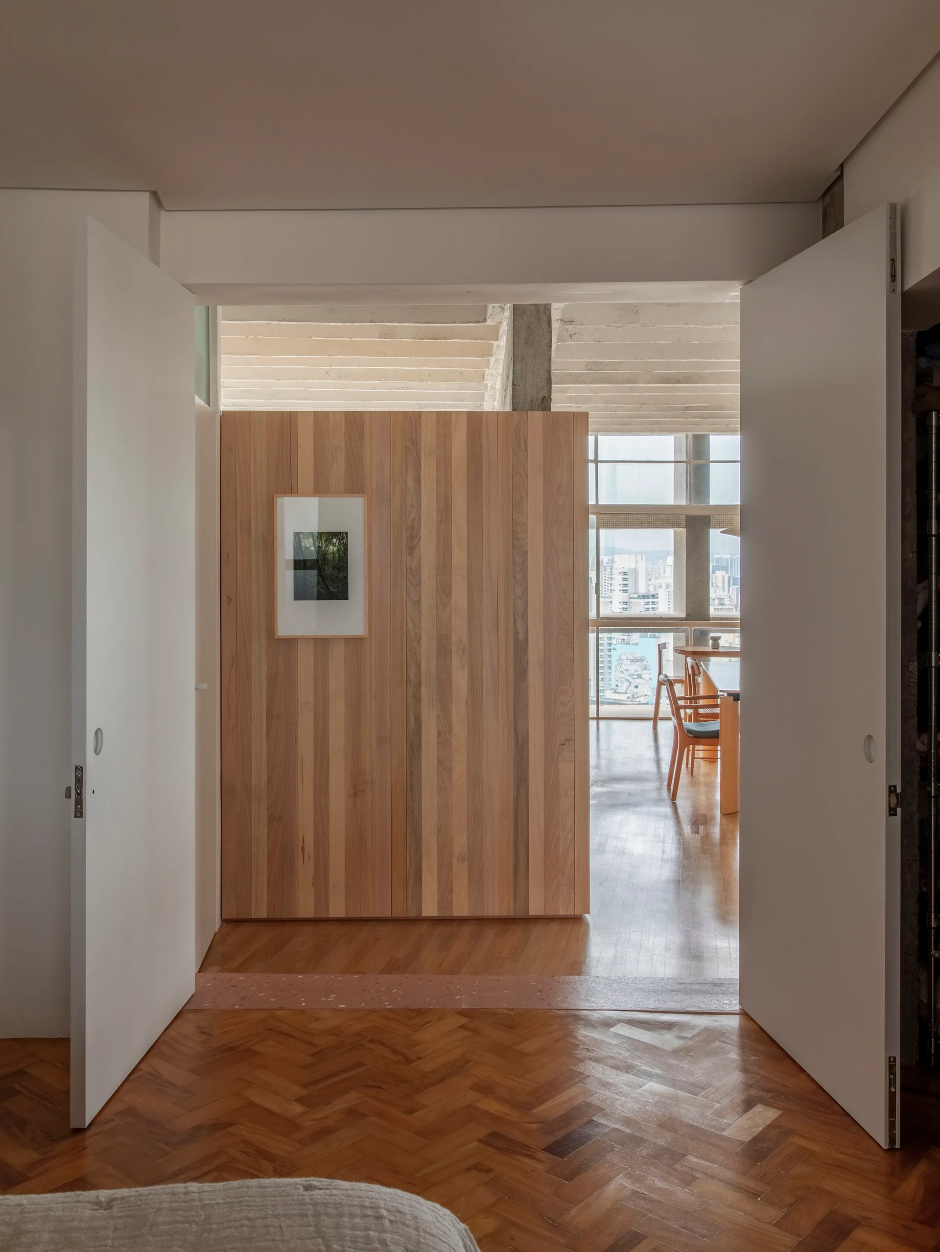 A view through open white doors reveals a vertical wood plank partition wall and a sunlit dining area with herringbone floors.