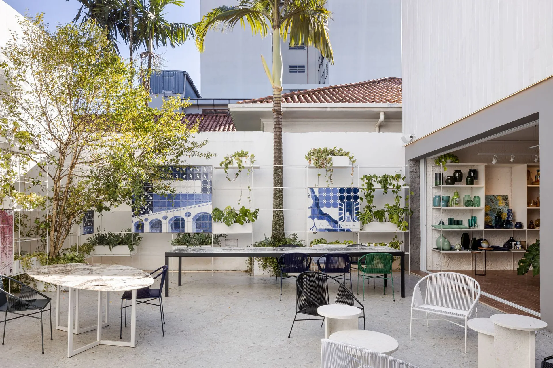 A sunny white courtyard features a long black dining table, assorted metal wire chairs, a palm tree, and blue tile art on metal shelves.