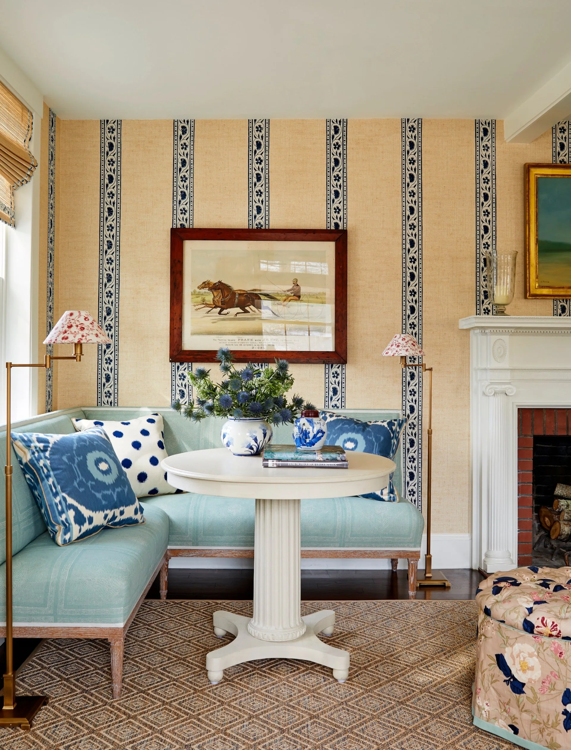 A light blue sectional sofa and a white pedestal table sit against tan and blue striped wallpaper near a white brick fireplace.