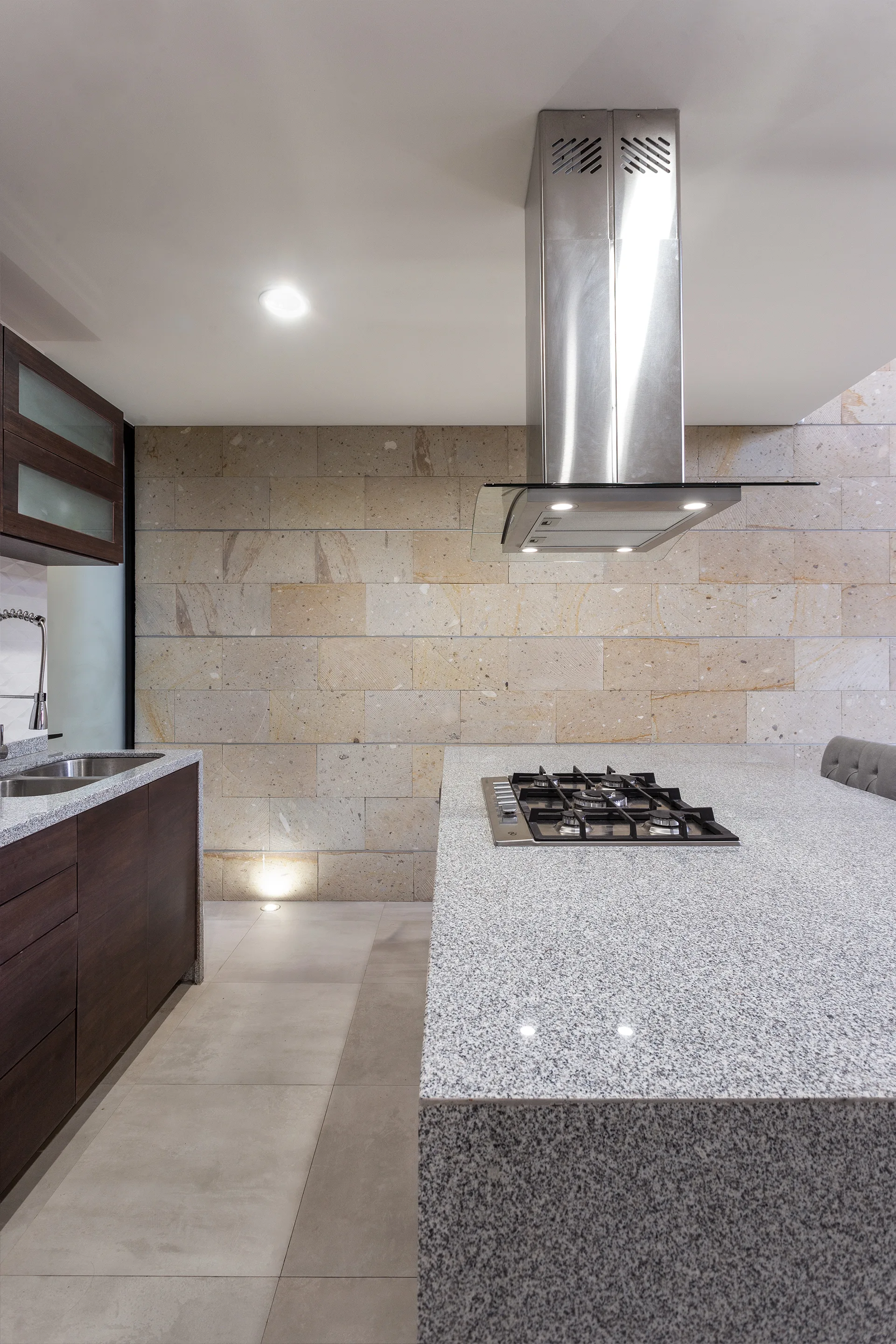 A modern kitchen showcasing a speckled granite island with a built-in cooktop, a stainless steel range hood, and a textured beige stone tile wall.