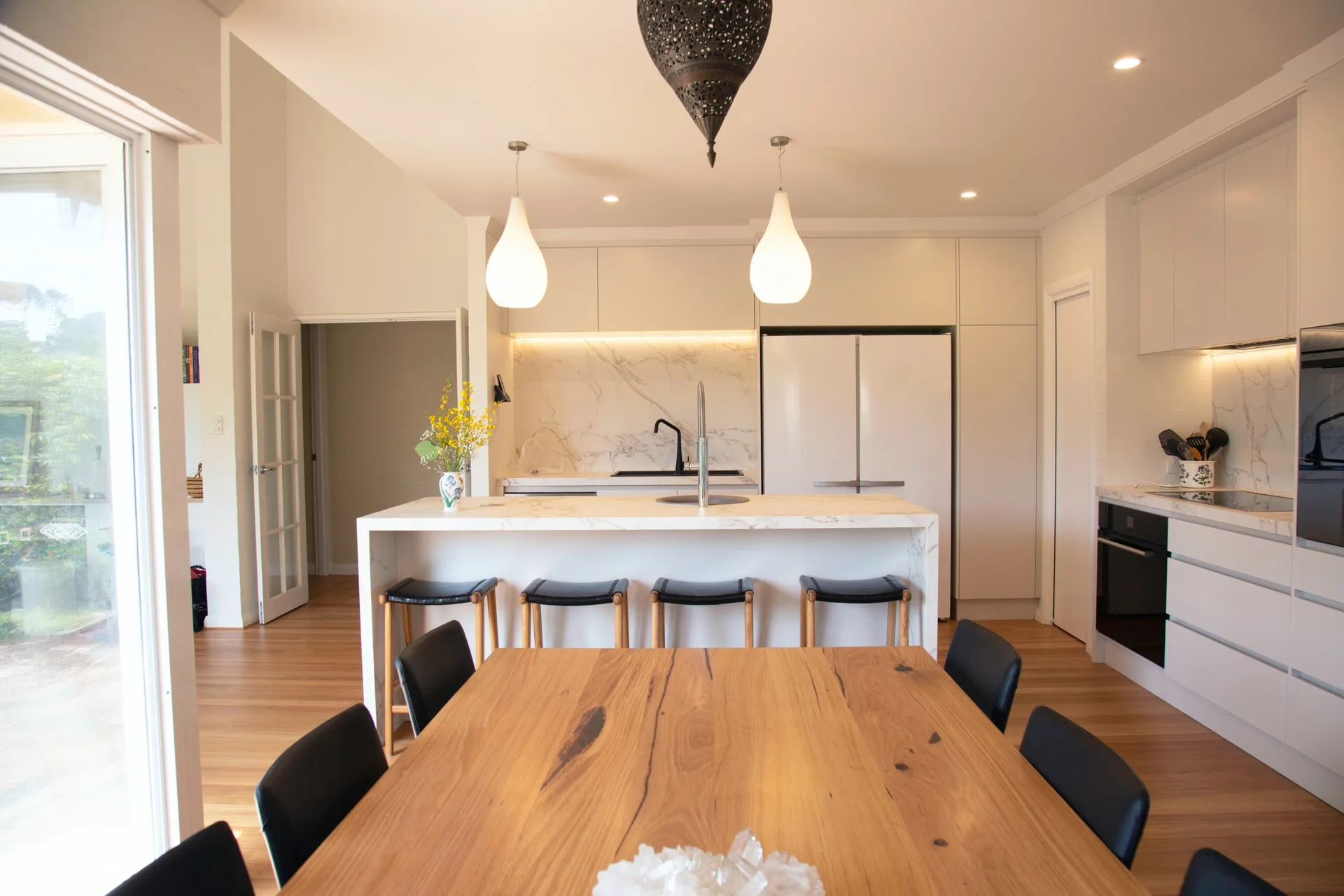 A modern kitchen featuring a marble waterfall island with four black stools positioned behind a natural wood dining table.