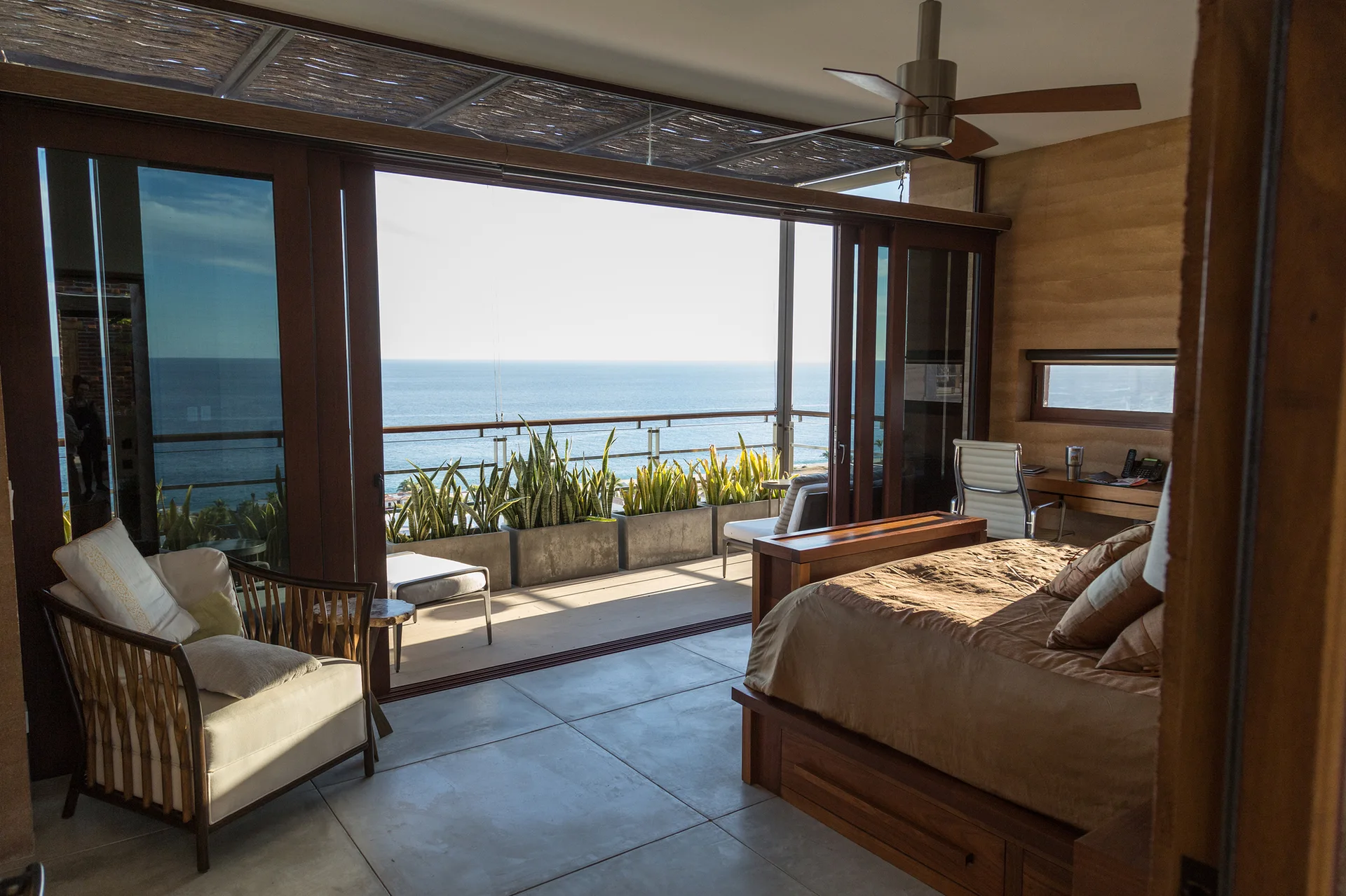 A modern bedroom featuring a wood platform bed, textured walls, and large sliding doors that open to a concrete tile terrace with ocean views.