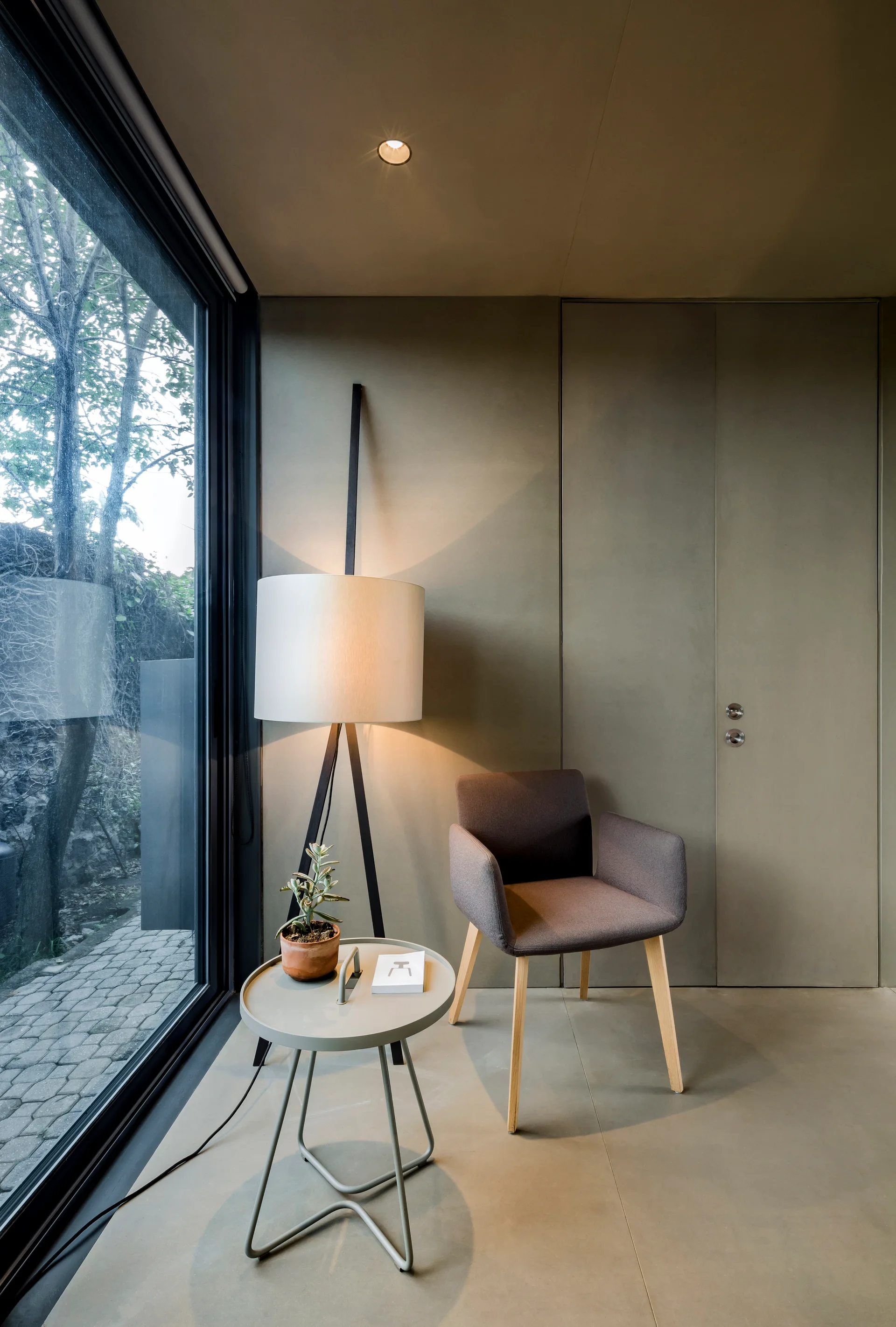 A minimalist room corner with a grey armchair, a tripod floor lamp, and a metal side table set against a taupe wall with an integrated flush door.