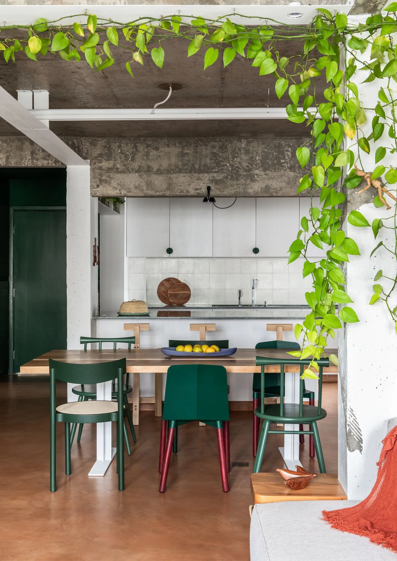 A modern dining space features a wooden table with green chairs below an exposed concrete ceiling and trailing green vines.